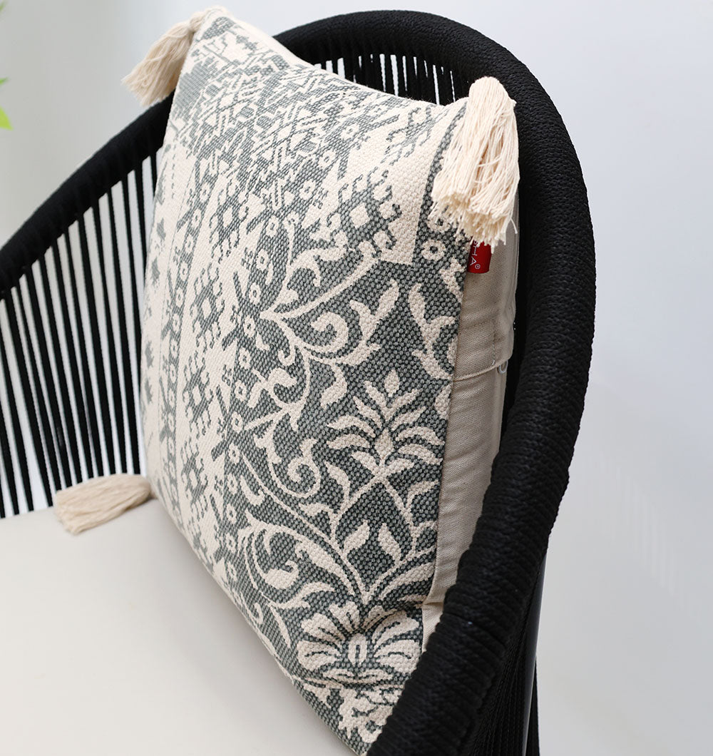 A black woven chair with a premium cotton cushion and a Ramsha Home White and Grey Printed Cotton Cushion 45x45 cm (CS 324), detailed with beige tassels on the corners.