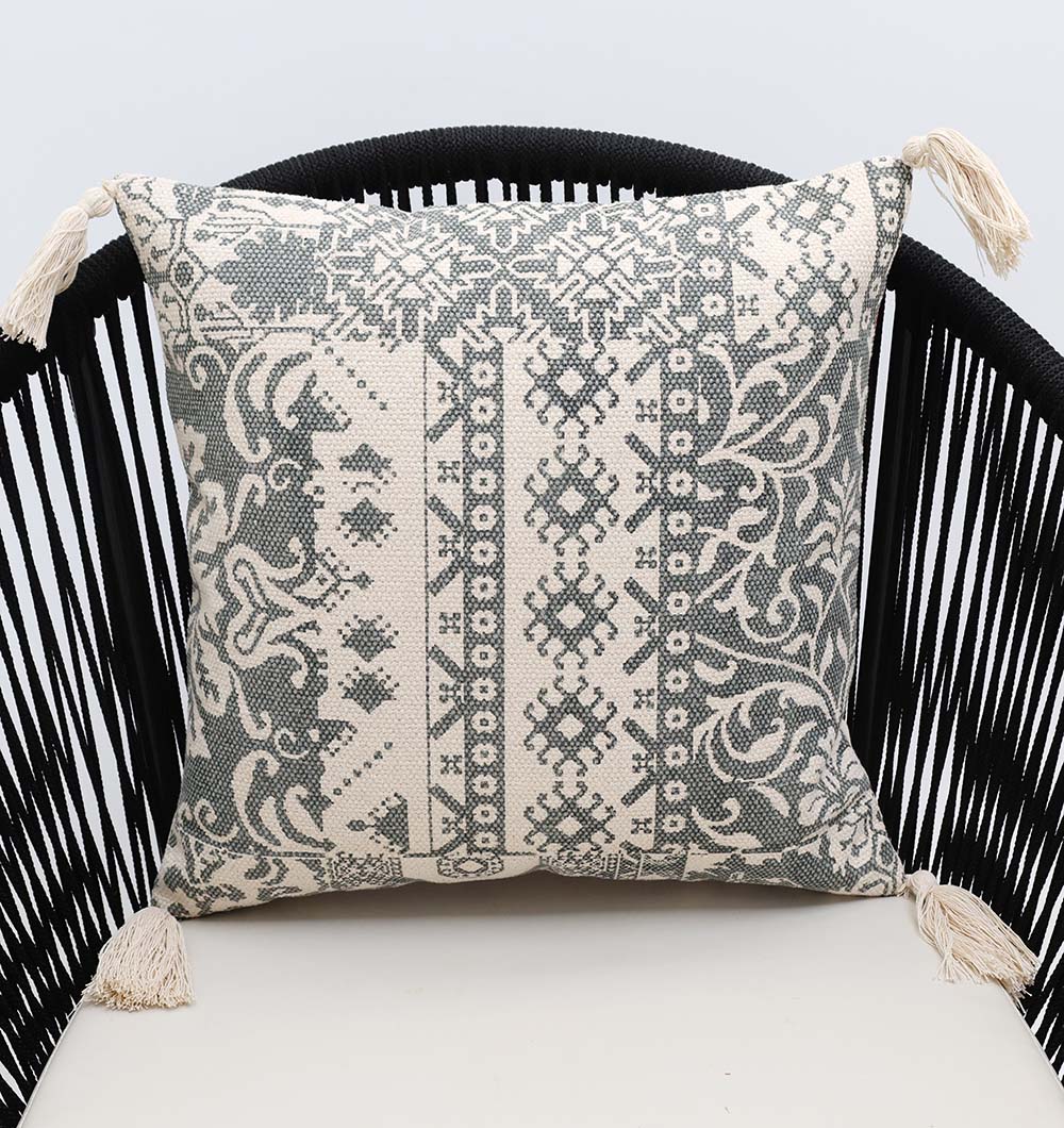 The Ramsha Home White and Grey Printed Cotton Cushion 45x45 cm (CS 324), featuring intricate geometric and floral designs with tassels, is elegantly placed on a black slatted curved-back chair.