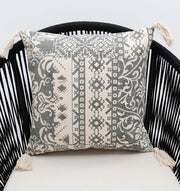 The Ramsha Home White and Grey Printed Cotton Cushion 45x45 cm (CS 324), featuring intricate geometric and floral designs with tassels, is elegantly placed on a black slatted curved-back chair.