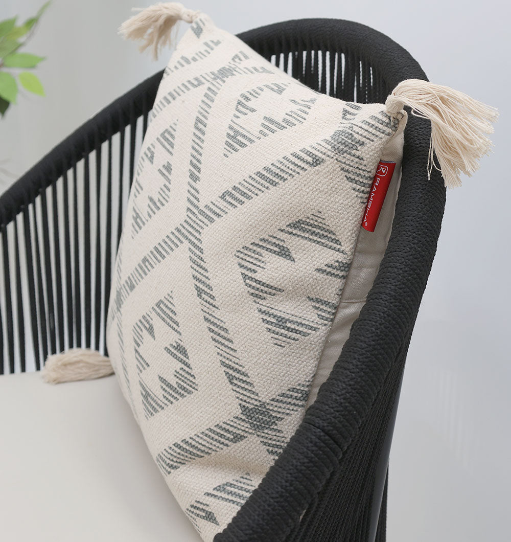 A black chair with woven sides holds the Ramsha Home Cream and Grey Printed Cotton Cushion 45x45 cm (CS 326). A small red tag is attached, and a green leafy plant appears in the background.