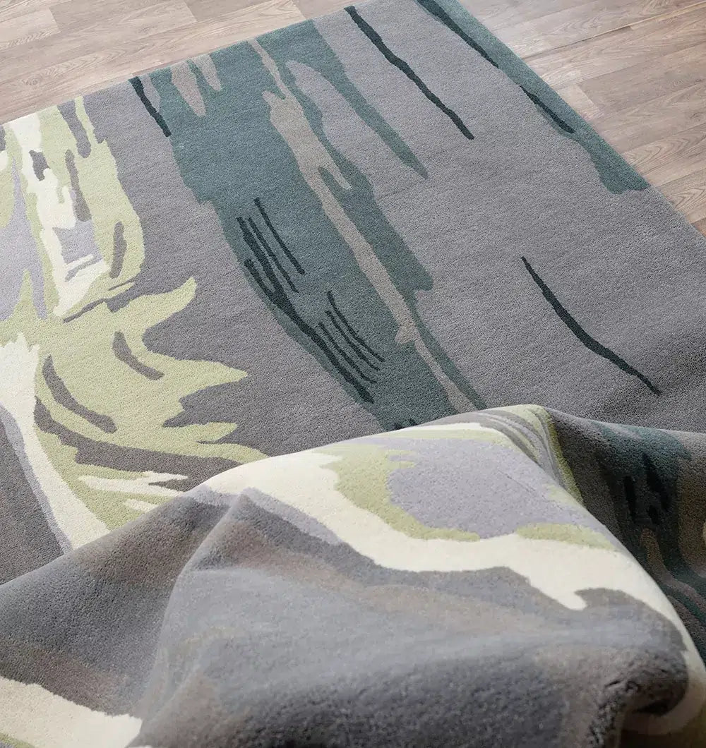 A close-up of the RAMSHA HOME Beautiful Hand-Tufted Rug - CWL 11 (200x300cm), in abstract gray, green, cream, and black tones, partially folded to showcase its texture and design on a wooden floor.