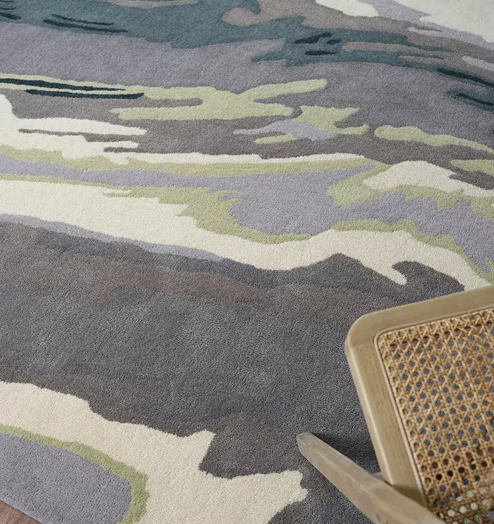 A close-up of the RAMSHA HOME Beautiful Hand-Tufted Rug - CWL 11 (200x300cm) in gray, green, and cream shades, displayed beside a wooden chair with a woven cane seat—a stylish accent for modern interiors.