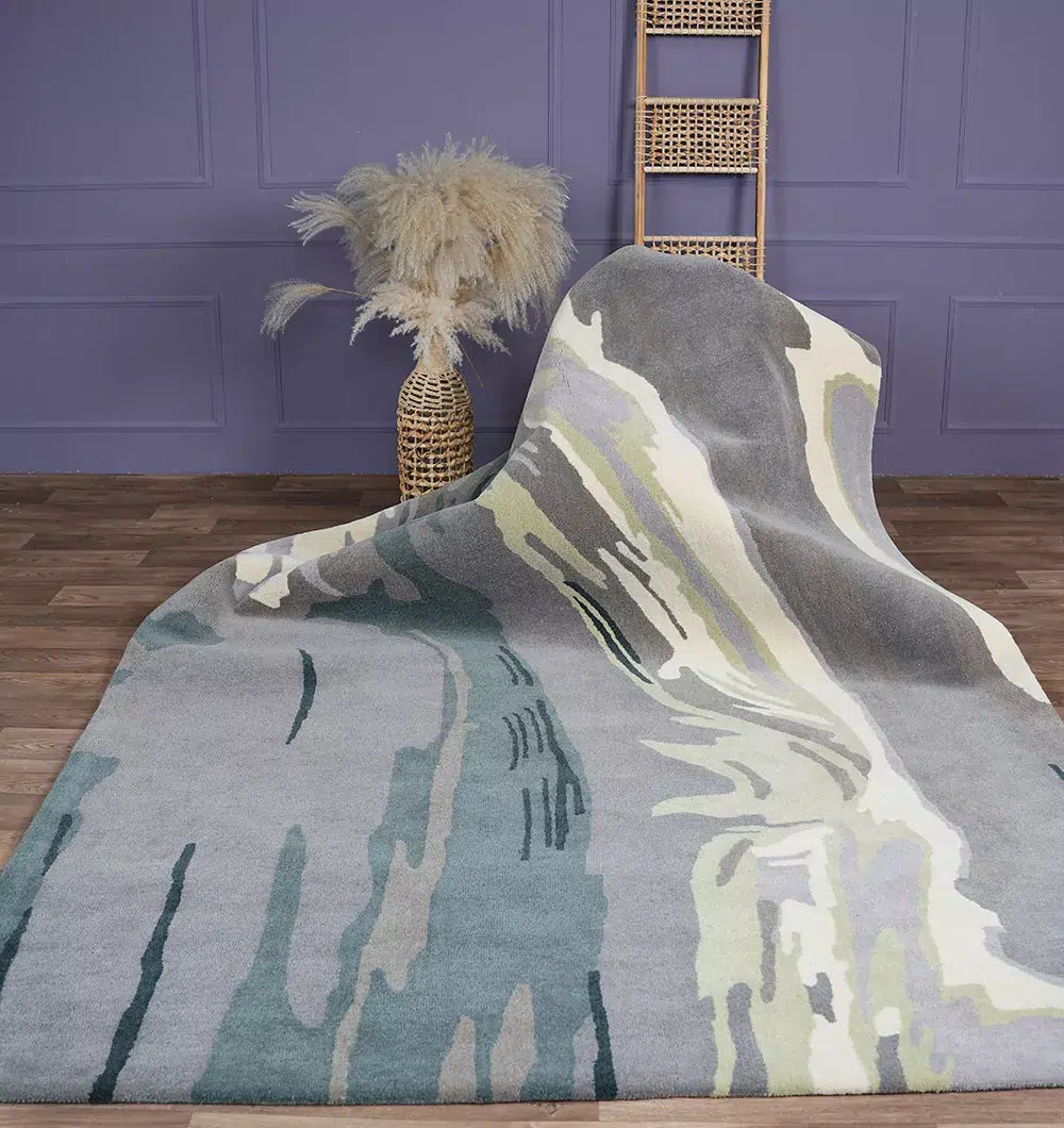 The RAMSHA HOME Beautiful Hand-Tufted Rug - CWL 11 (200x300cm) in blue-grey with abstract blue, green, and cream patterns is displayed on a wooden floor, styled with a vase of pampas grass and a bamboo ladder against a purple paneled wall.