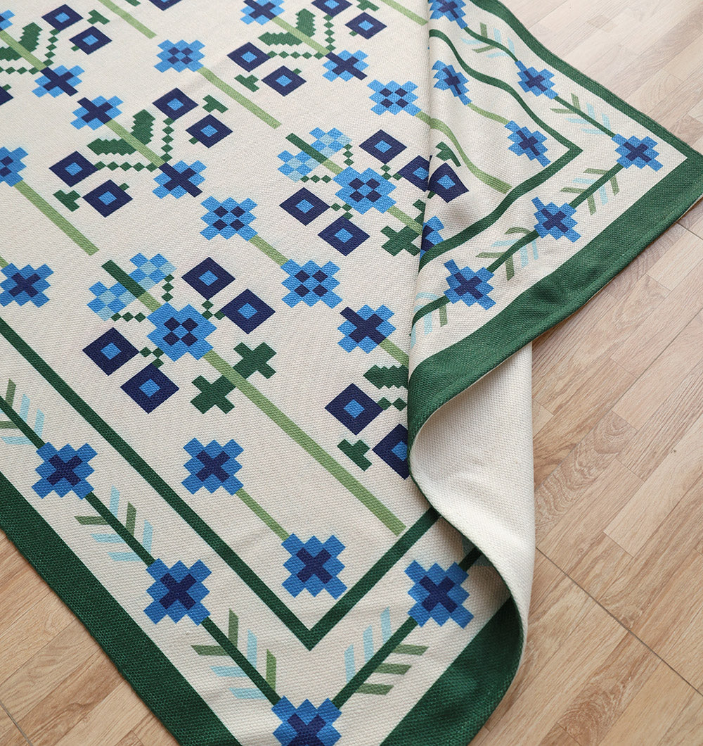 The RAMSHA HOME Blue Traditional Floral Digital Printed Rug – Bohemian Area Rug DD-049, featuring a pixelated blue and green floral pattern with a green border, is partially folded over on a light wood floor.