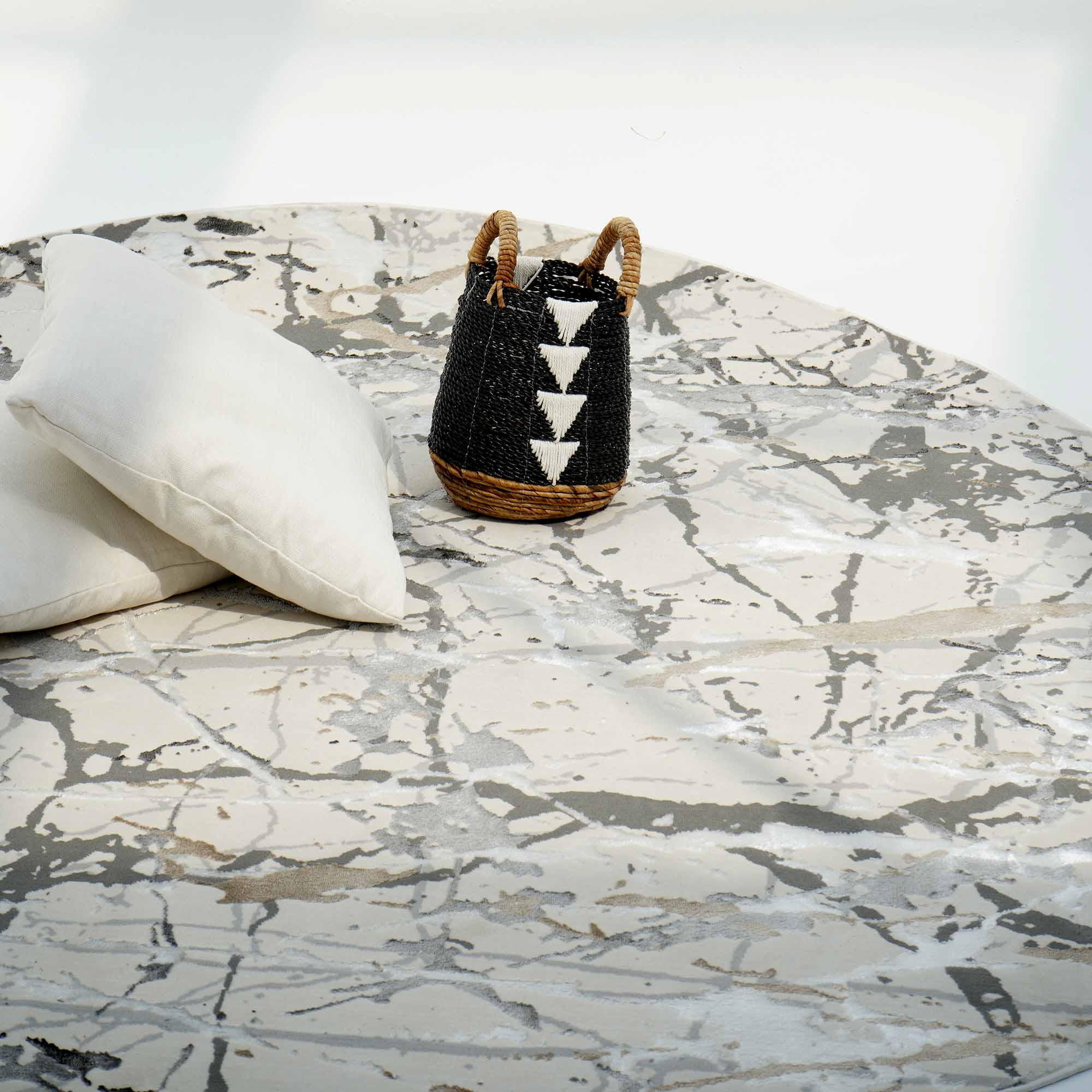 A round table with a white and gray abstract top, two white pillows, and a small black basket sits atop the RAMSHA HOME Machine Made Turkey Carpet Cream Colored MZ29C.