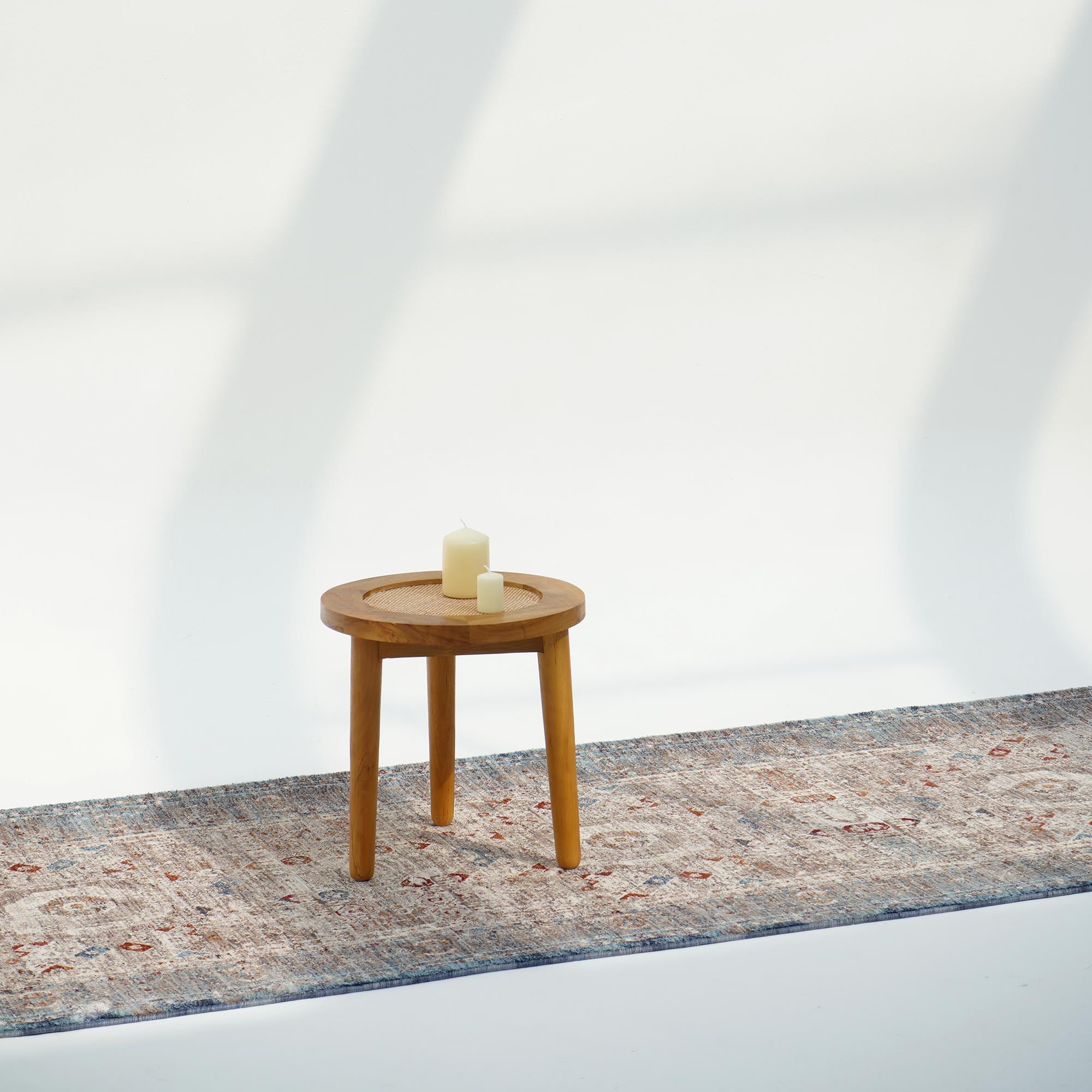A small round wooden table with two pillar candles on top stands on a RAMSHA HOME Polyester K860A Area Rug, set against a plain white background with soft shadows.