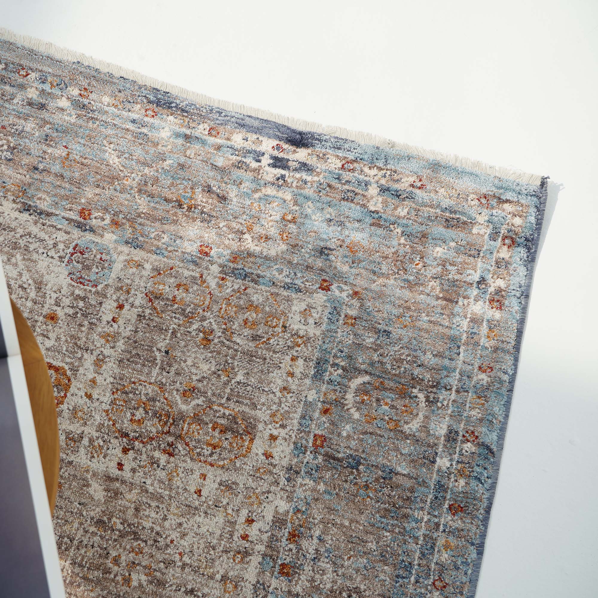 A close-up of the RAMSHA HOME Polyester K860A Area Rug, featuring blue, beige, and brown tones with a distressed look, is slightly folded at one corner on a white floor in natural light.