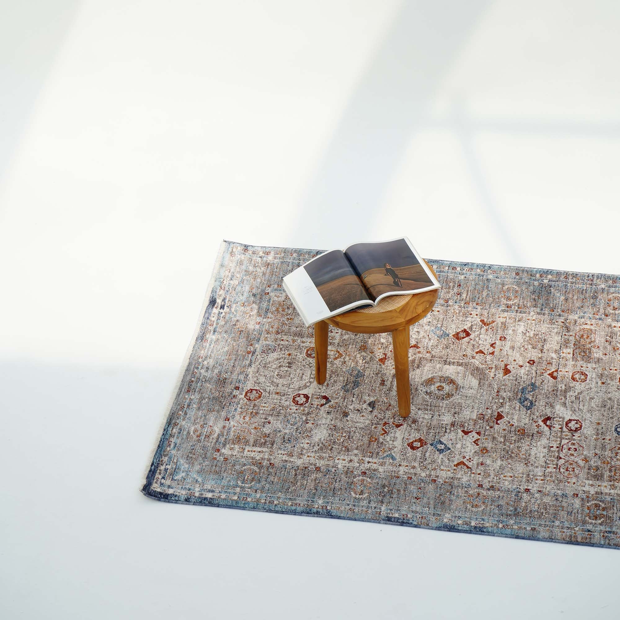 A small wooden stool with an open book sits on the RAMSHA HOME Polyester K860A Area Rug, featuring blue and red designs, set against a white background with soft shadows.
