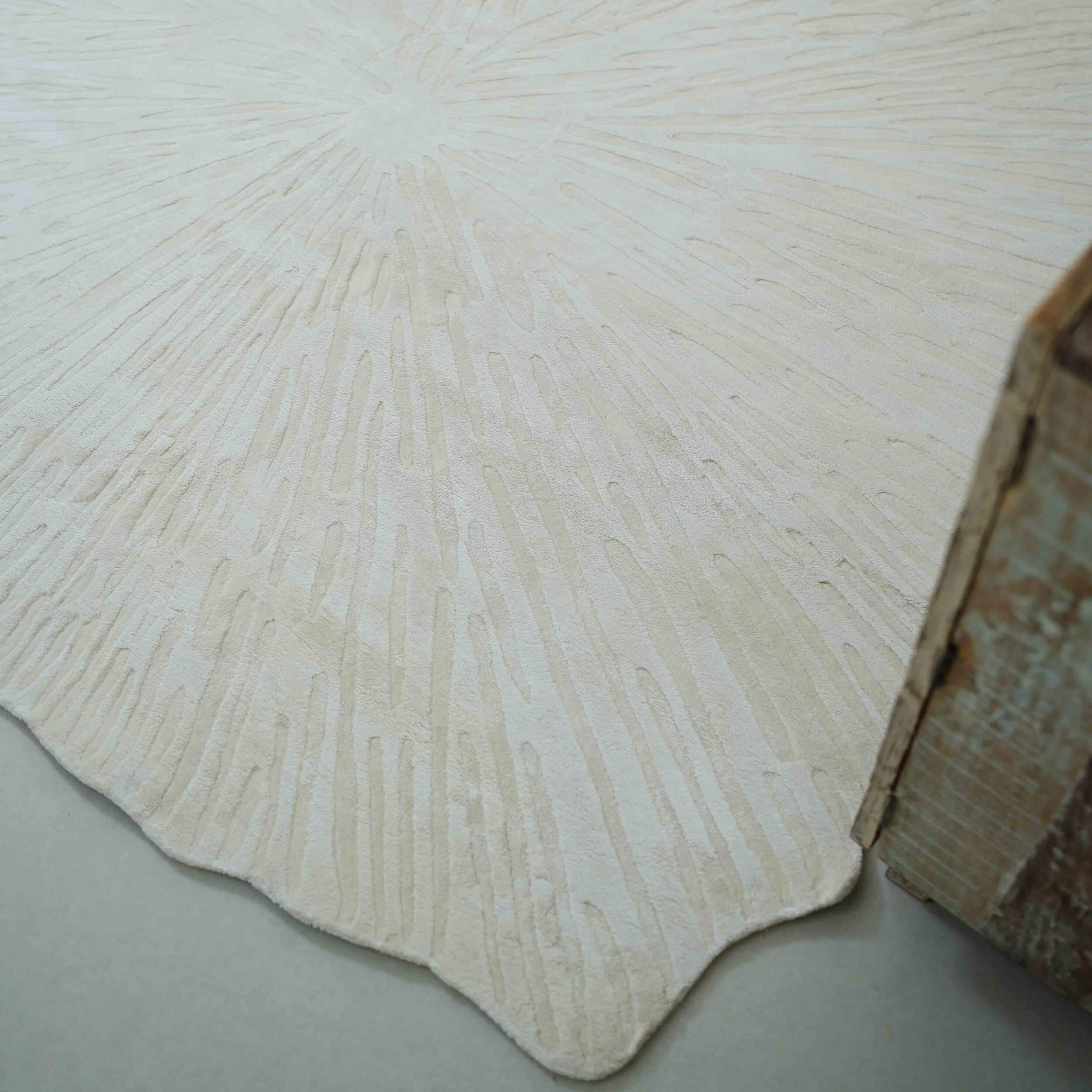 A close-up of the RAMSHA HOME Home Decor Shape Handmade Area Rug - HTV 21, featuring textured ivory fabric with irregular radiating lines and a wavy edge, is shown beside the corner of a wooden furniture piece.
