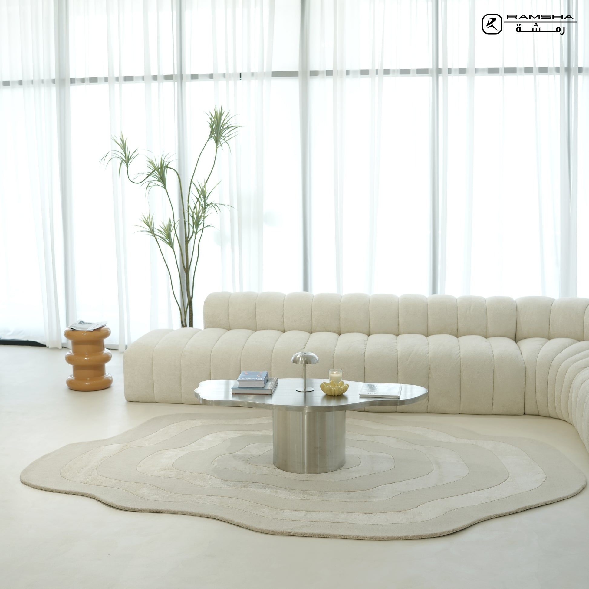 A modern, minimalist living room with a curved beige sofa, RAMSHA HOME’s Wool & Tencel Multi Color Rug HTWV-91 featuring wavy patterns, a silver coffee table, a table lamp, books, a tall plant, wooden side table, and sheer white curtains letting in natural light.