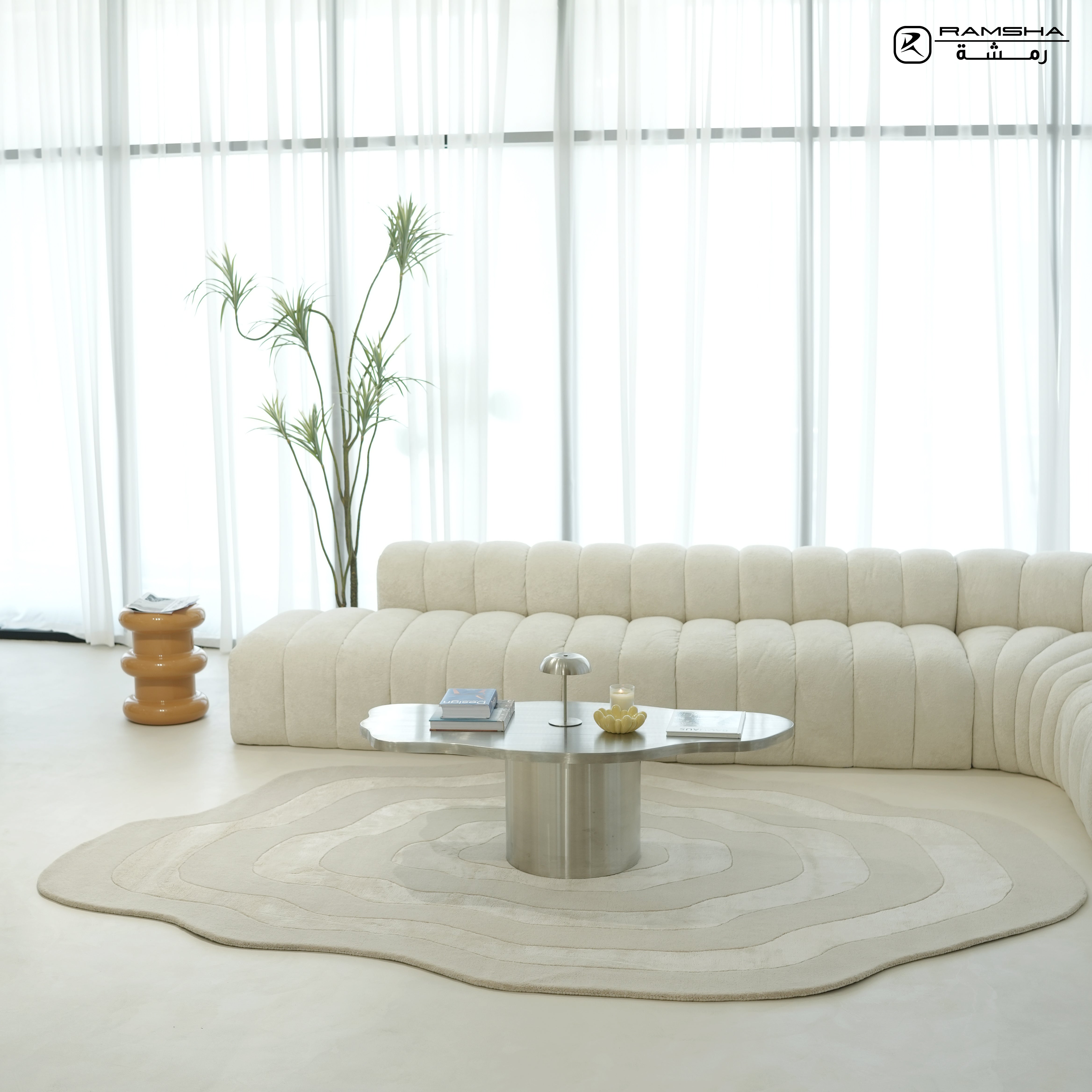 A modern, minimalist living room with a curved beige sofa, RAMSHA HOME’s Wool & Tencel Multi Color Rug HTWV-91 featuring wavy patterns, a silver coffee table, a table lamp, books, a tall plant, wooden side table, and sheer white curtains letting in natural light.