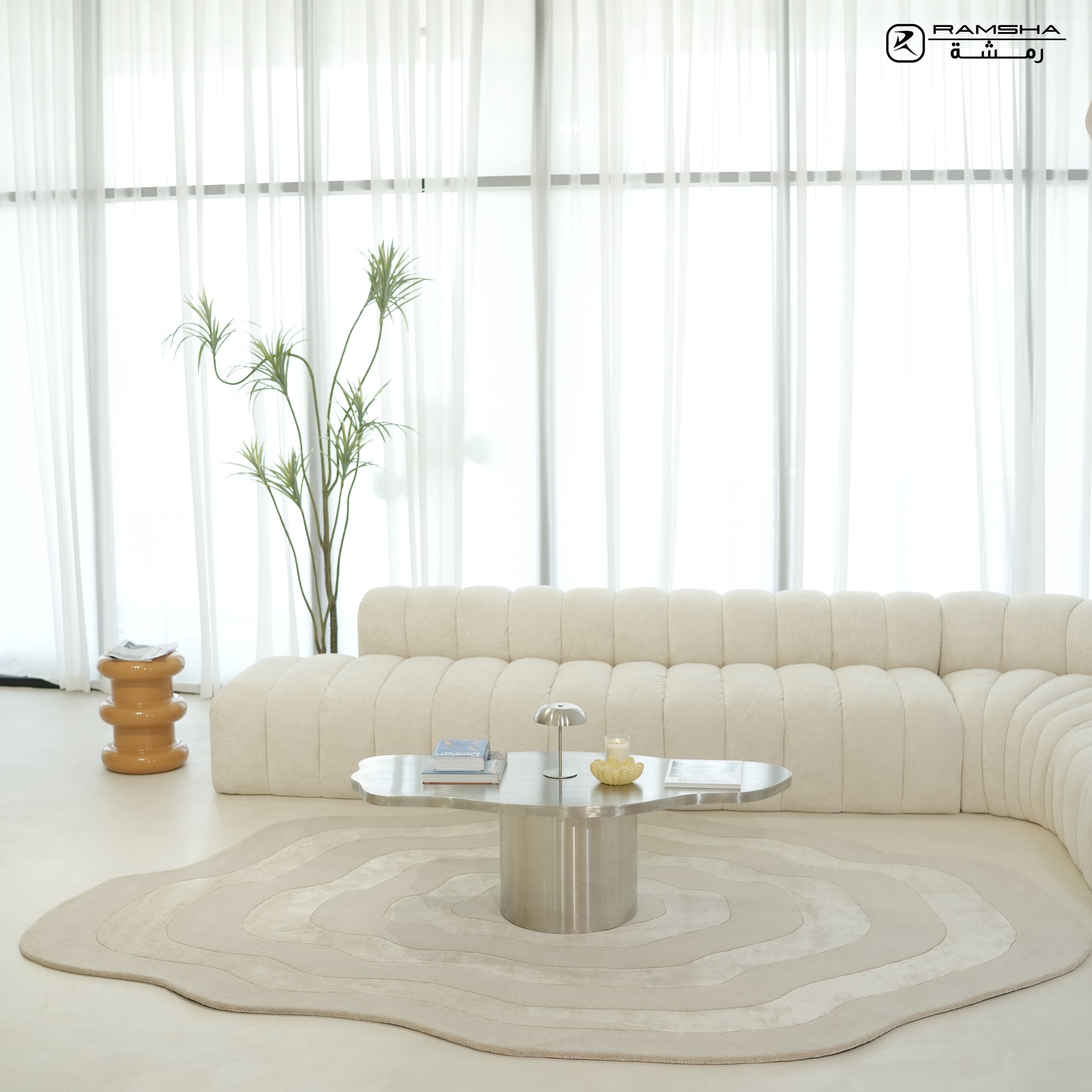 A modern, minimalist living room featuring a curved cream sofa, RAMSHA HOME's Wool & Tencel Multi Color Rug HTWV-91, a metallic coffee table with books and decor, a tall potted plant, and sheer white curtains streaming in natural light.