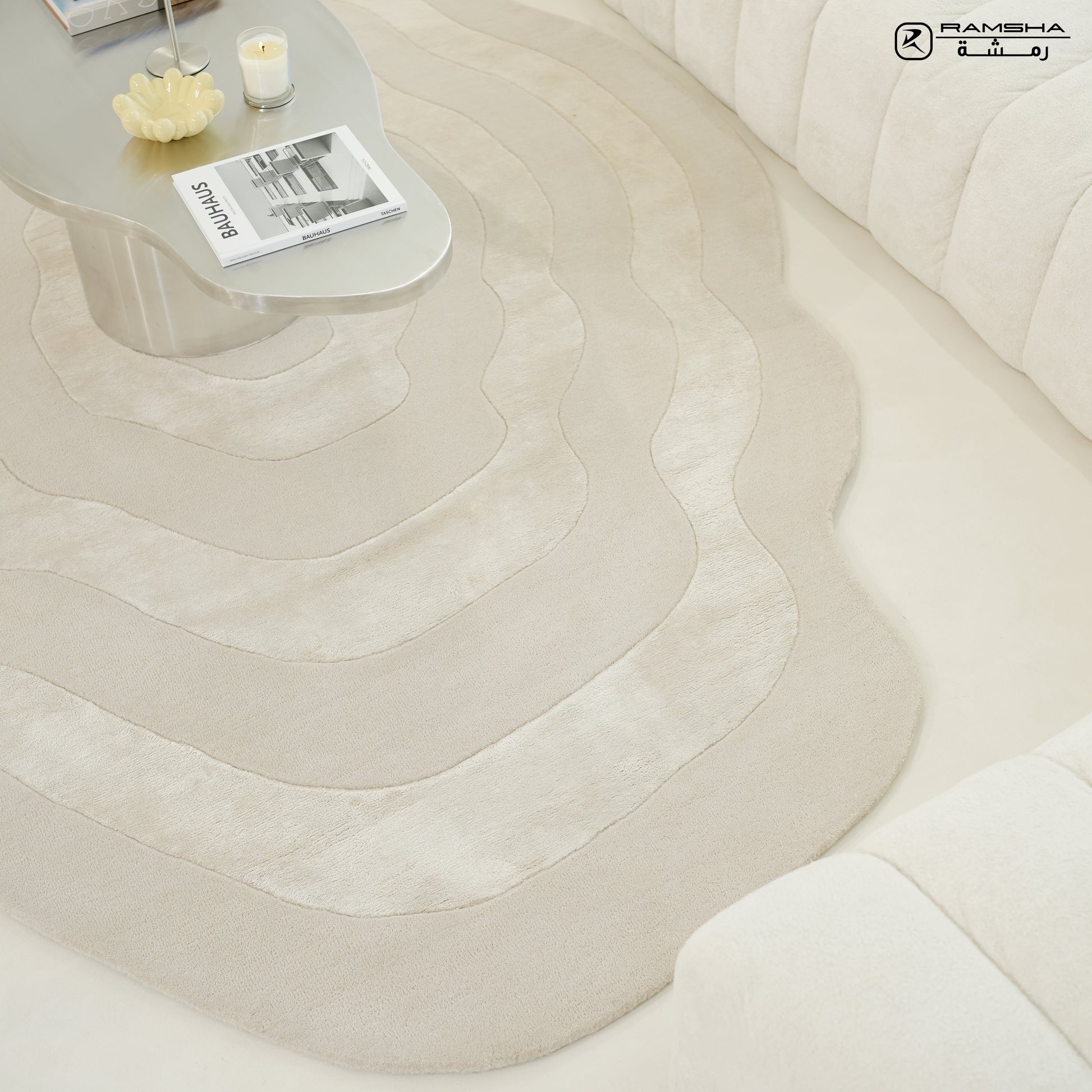 A bright, minimalist living room features the RAMSHA HOME Wool & Tencel Multi Color Rug HTWV-91, paired with a curved white sofa and a sleek pale coffee table displaying a candle and magazines.