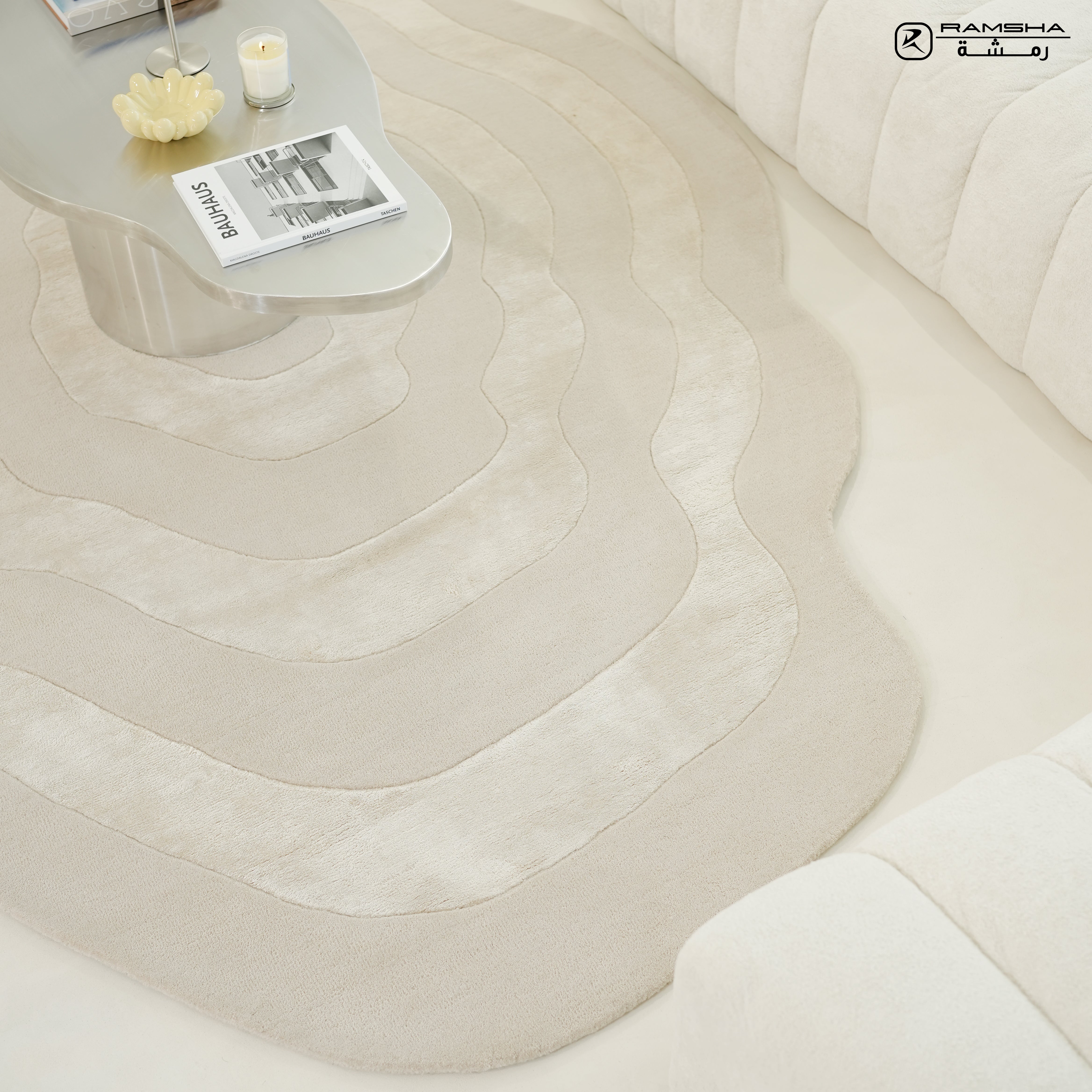 A bright, minimalist living room features the RAMSHA HOME Wool & Tencel Multi Color Rug HTWV-91, paired with a curved white sofa and a sleek pale coffee table displaying a candle and magazines.