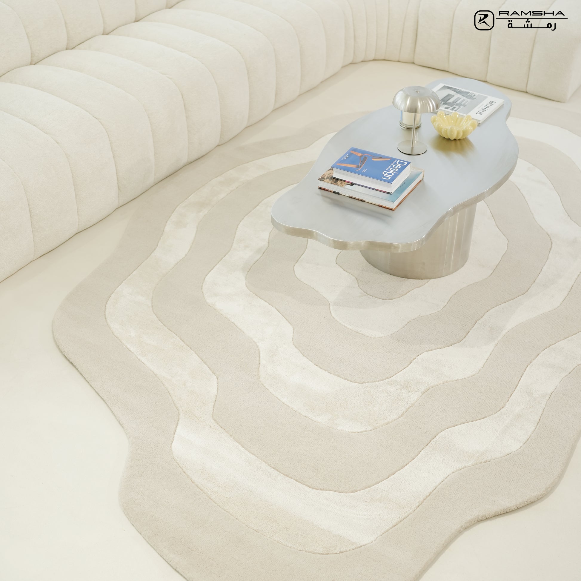 A modern living room features a curved cream sofa, a RAMSHA HOME Wool & Tencel Multi Color Rug HTWV-91 in beige tones, and a silver sculptural coffee table with books, a lamp, and décor. The space feels minimal and elegant.