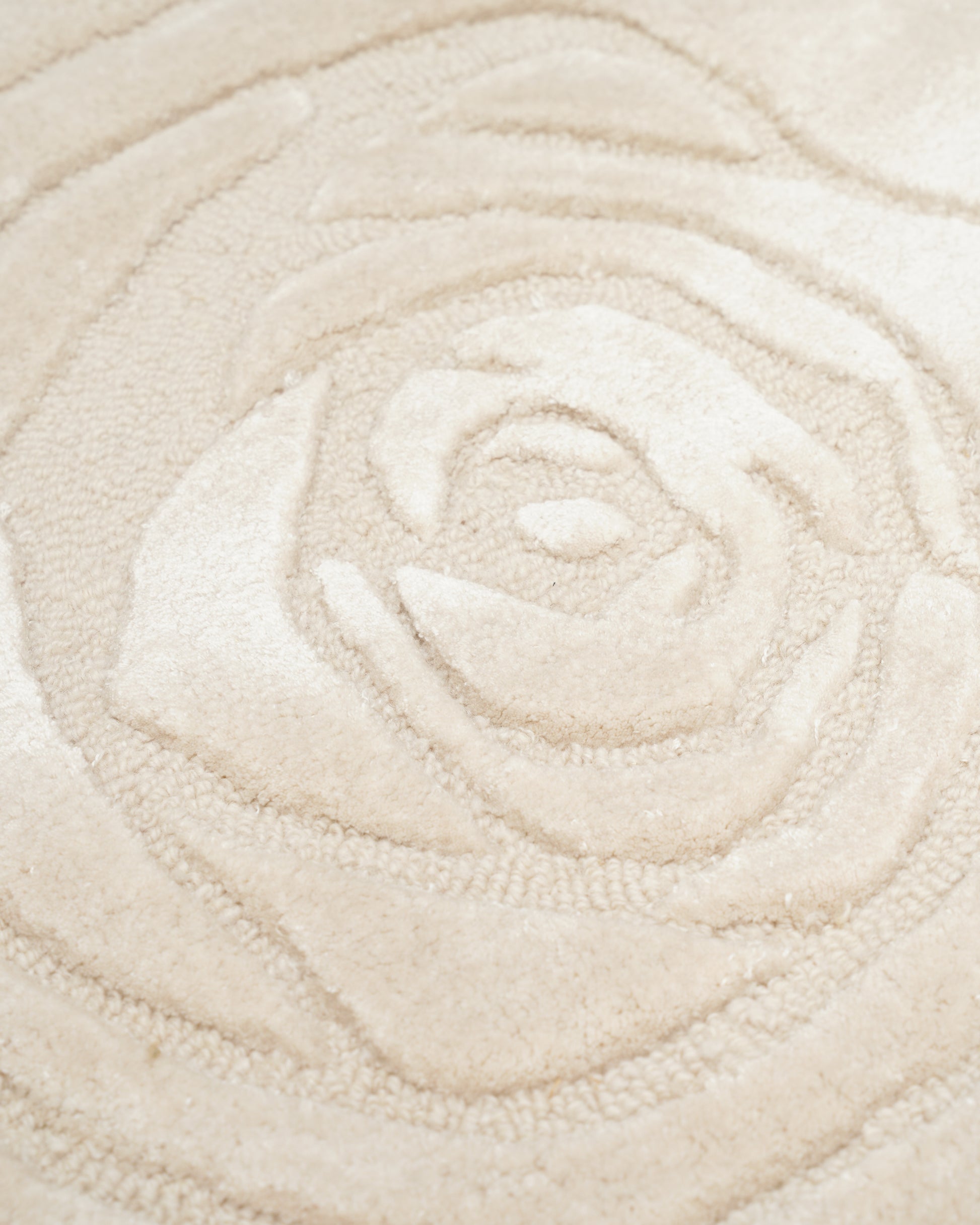 A close-up of the RAMSHA HOME Home Decor Handmade Area Rug - HTWV-156 in beige showcases its raised, carved floral pattern reminiscent of a rose, enhanced by soft lighting. The textured design adds subtle depth and dimension, accentuating the rug’s elegant craftsmanship.
