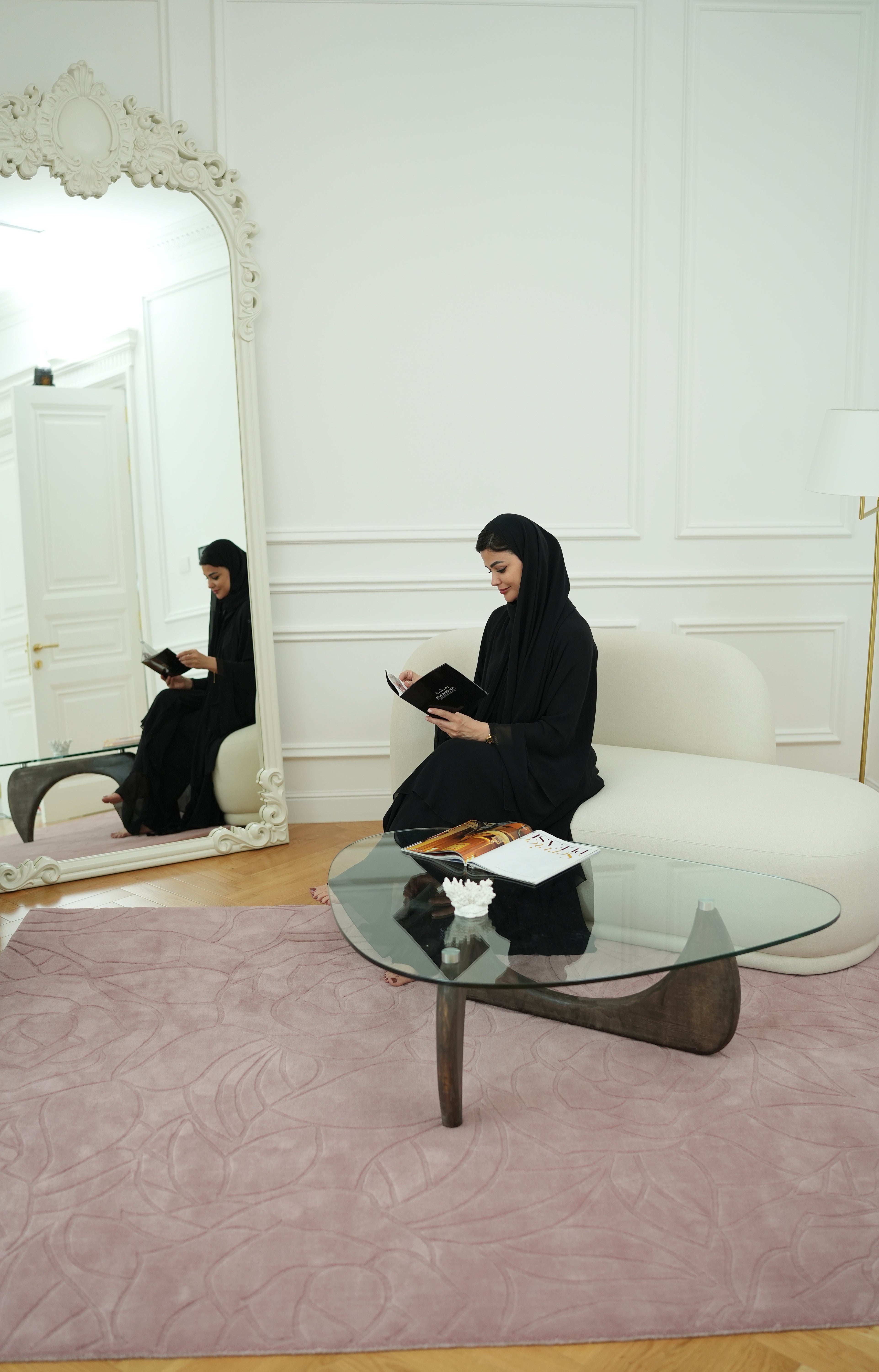 A woman in a black abaya sits on a white sofa reading a book in an elegant room, featuring a glass coffee table and the RAMSHA HOME Rectangle Handmade Pink Color Area Rug (LKH-029); her reflection appears in the large ornate mirror.