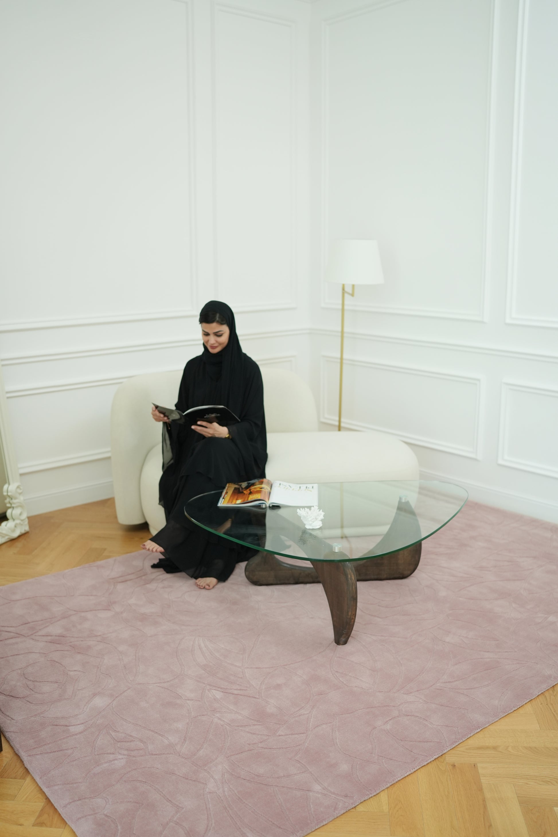 A woman in a black abaya sits on a white sofa, reading a book in a bright room with white walls, a glass coffee table, and the RAMSHA HOME Rectangle Handmade Pink Color Area Rug (LKH-029). A lamp stands in the corner.