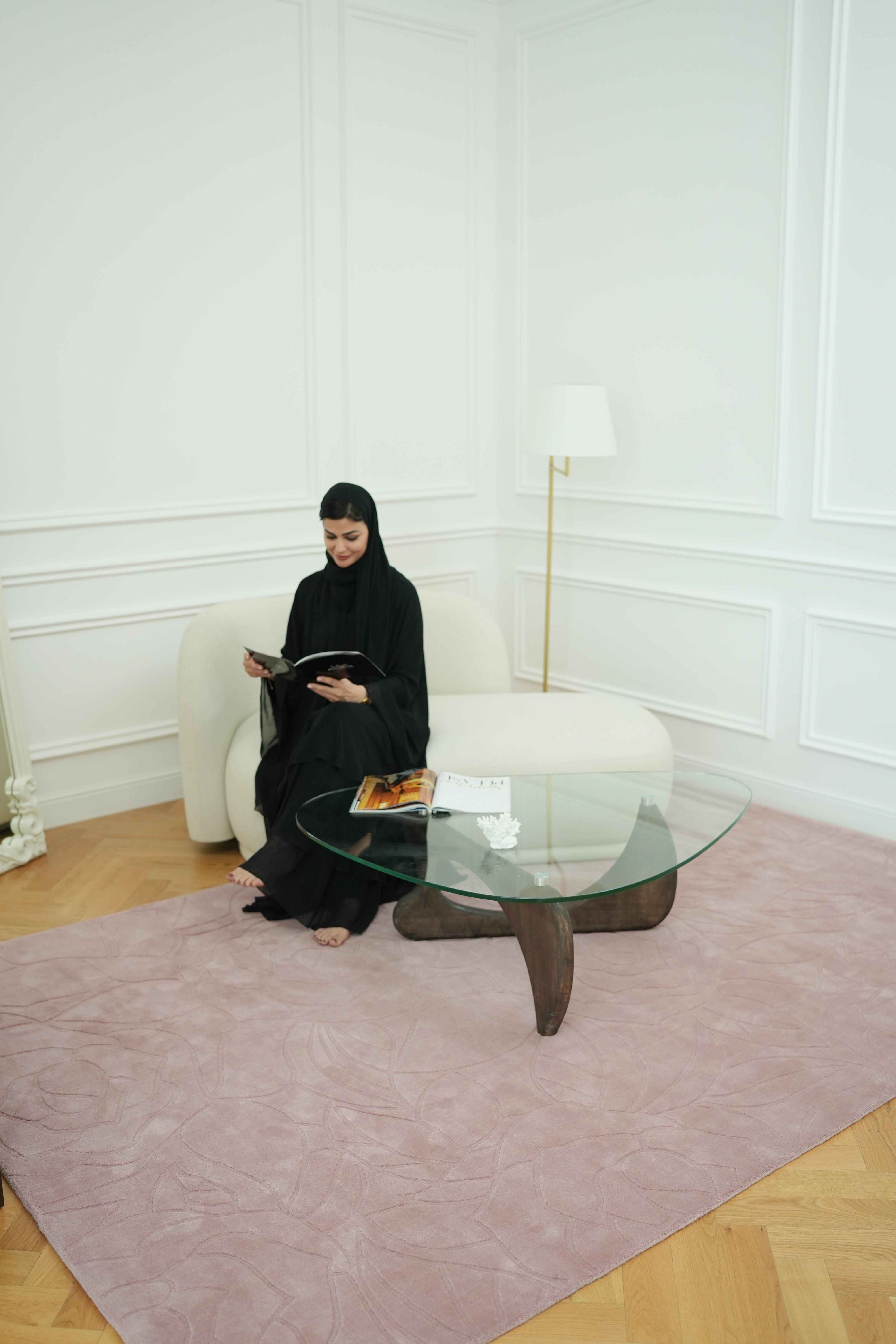 A woman in a black abaya sits on a white sofa, reading a book in a bright room with white walls, a glass coffee table, and the RAMSHA HOME Rectangle Handmade Pink Color Area Rug (LKH-029). A lamp stands in the corner.