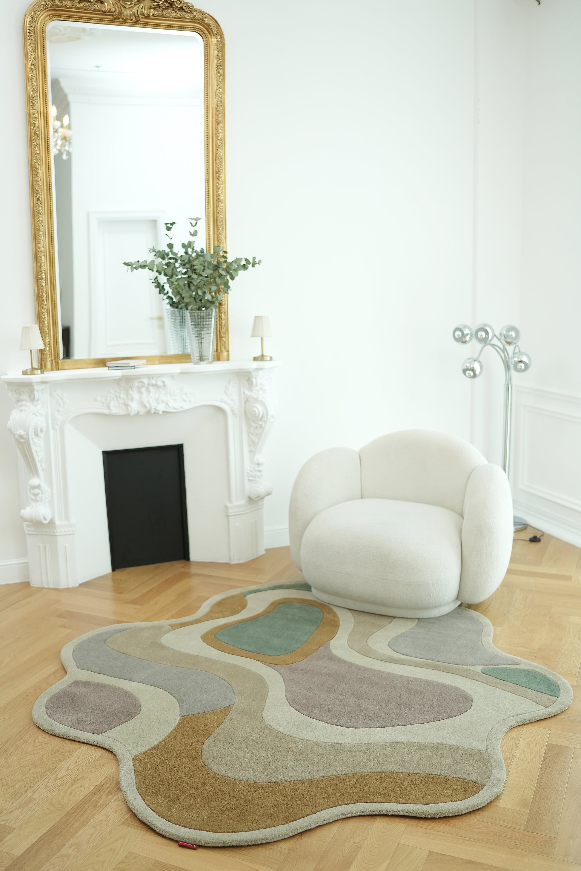 A modern, minimalist room with a white armchair, the RAMSHA HOME Handmade Multicolor Shaped Area Rug HTW 127 (200cm), a white ornate fireplace topped by a gold-framed mirror, greenery in a vase, and a metallic floor lamp in the corner.