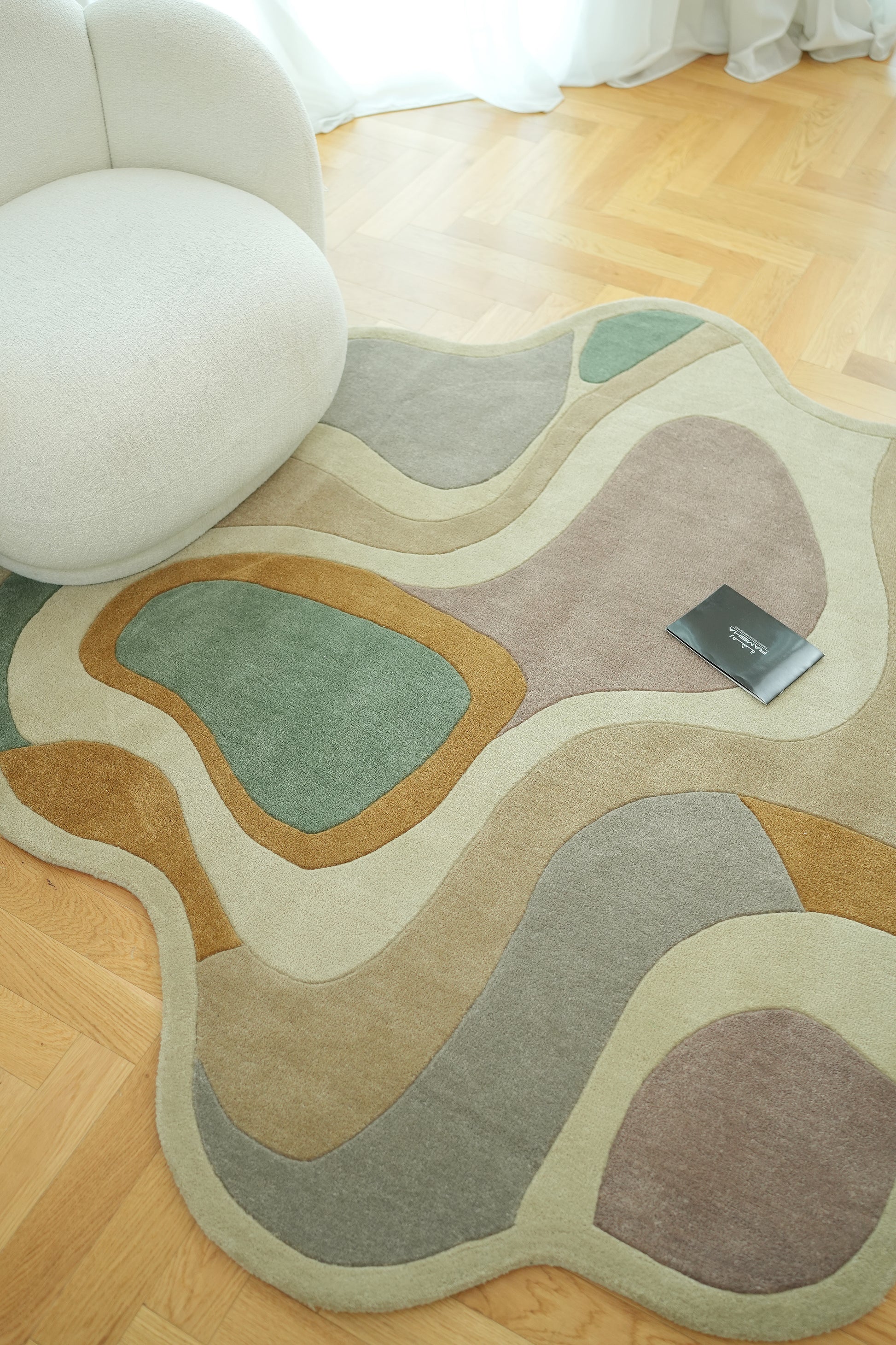 The RAMSHA HOME Handmade Multicolor Shaped Area Rug HTW 127 (200cm), featuring curved green, tan, brown, and beige designs, lies on a wooden floor by a rounded cream chair with a black booklet resting on it.