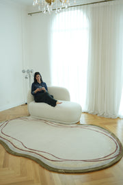A woman sits reading on a white chaise lounge in a bright room with large windows, sheer curtains, wooden floors, and the RAMSHA HOME Handmade Hand-Tufted Carpet HTWV-60.