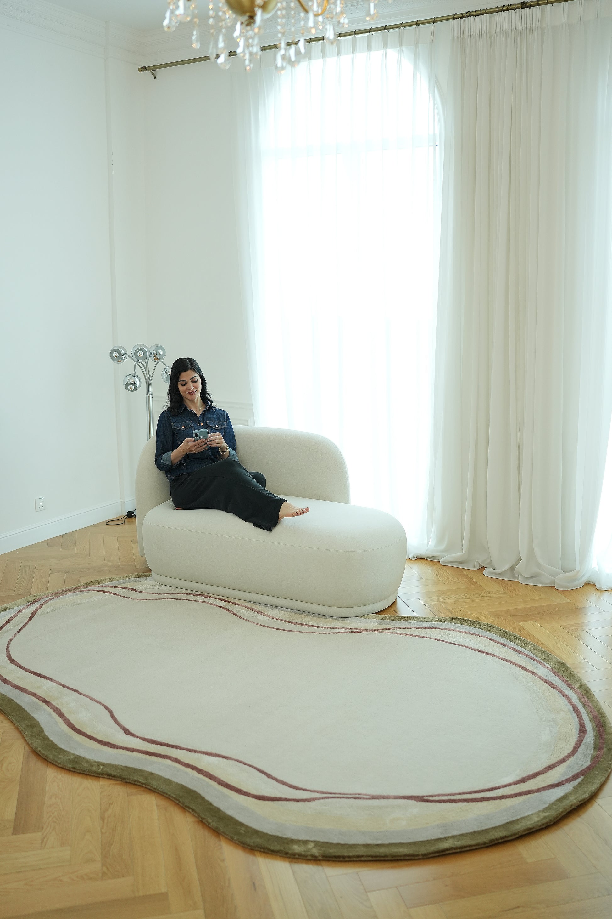 A woman sits reading on a white chaise lounge in a bright room with large windows, sheer curtains, wooden floors, and the RAMSHA HOME Handmade Hand-Tufted Carpet HTWV-60.