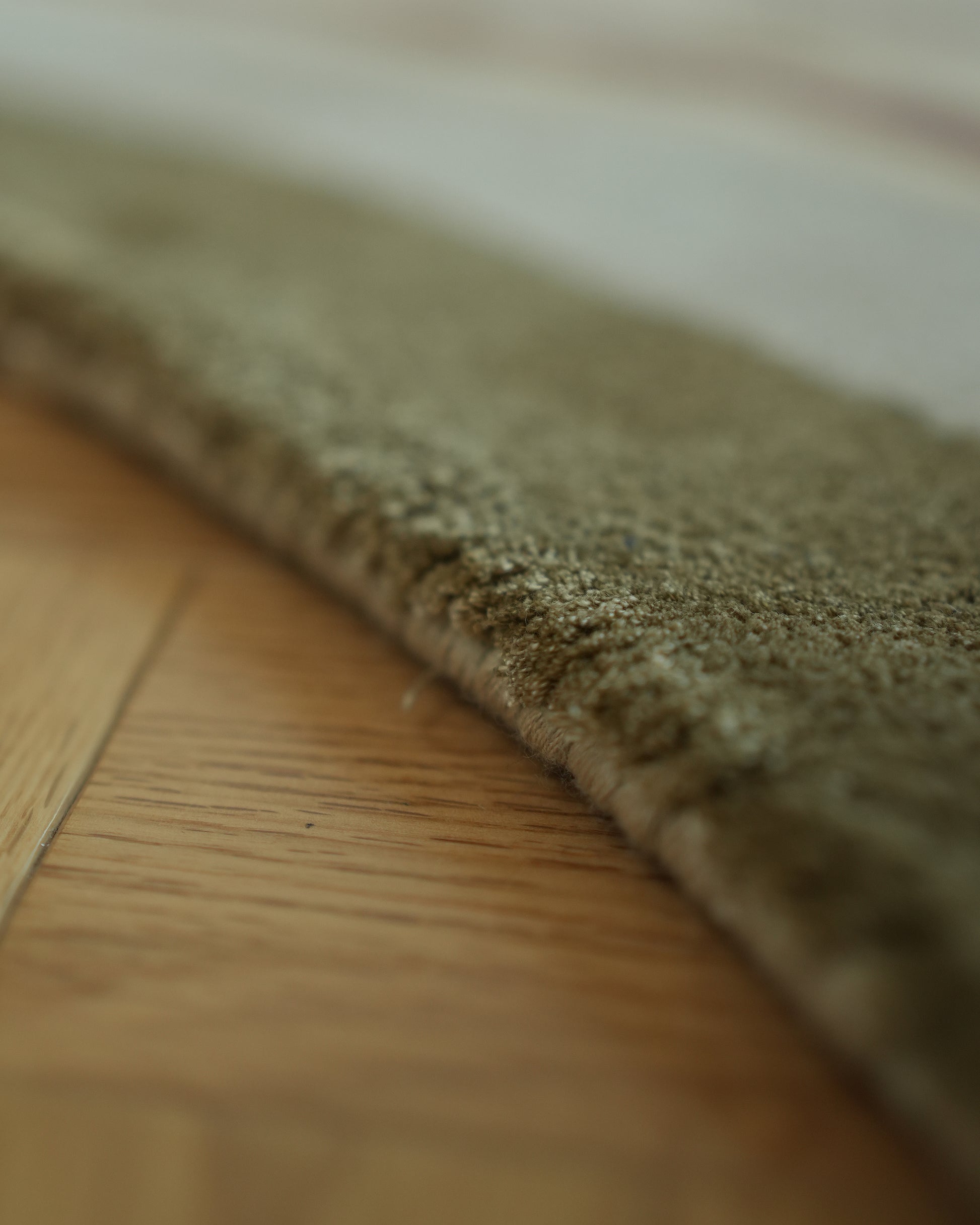 Close-up of the Handmade Hand-Tufted Carpet HTWV-60 by RAMSHA HOME, showing its plush greenish-brown fibers against a wooden floor, highlighting the texture and striking contrast with the smooth wood surface.