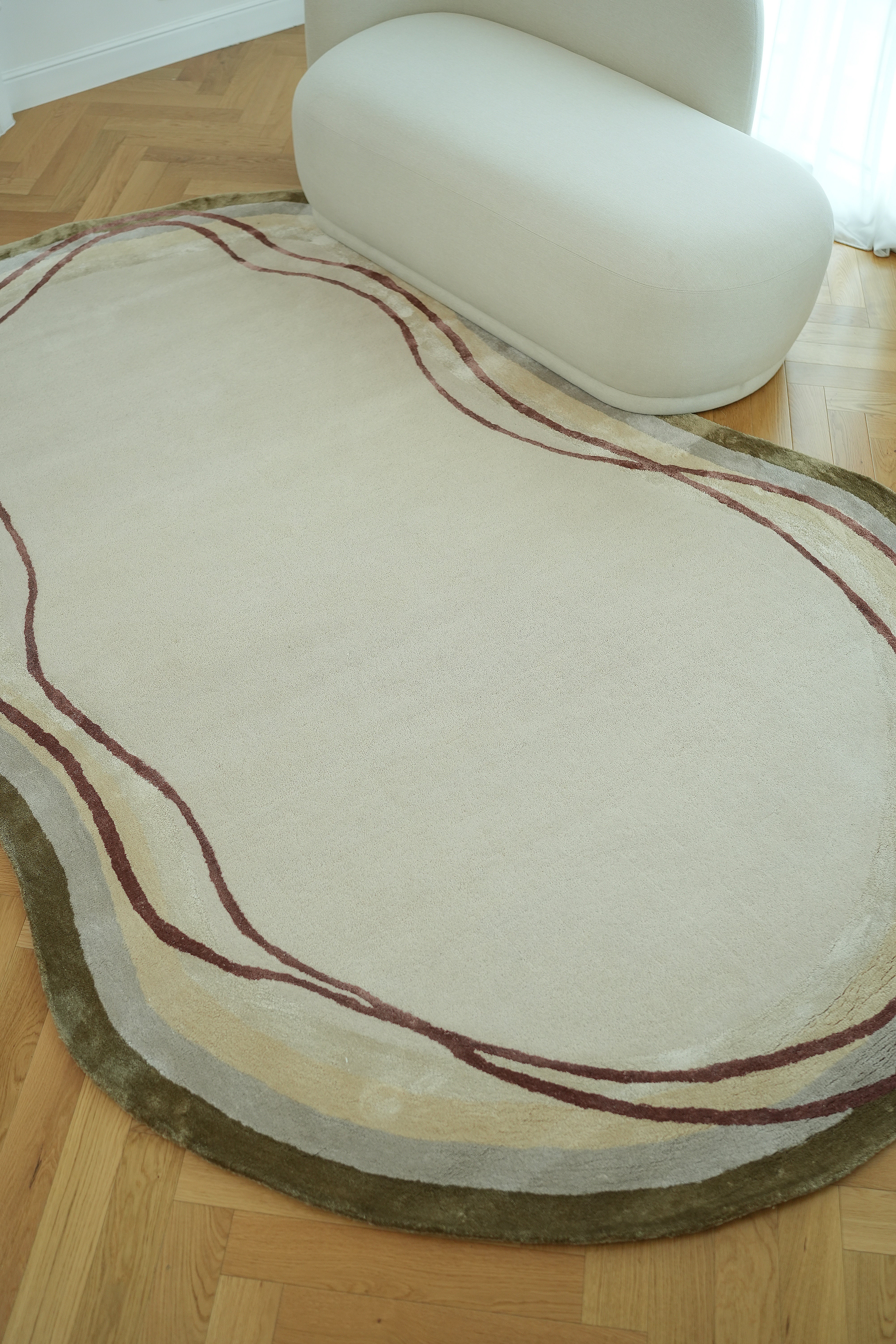 The RAMSHA HOME Handmade Hand-Tufted Carpet HTWV-60, featuring an abstract beige design with wavy red and green lines, is shown on a wooden floor beside a minimalist cream curved sofa near a window with sheer white curtains.