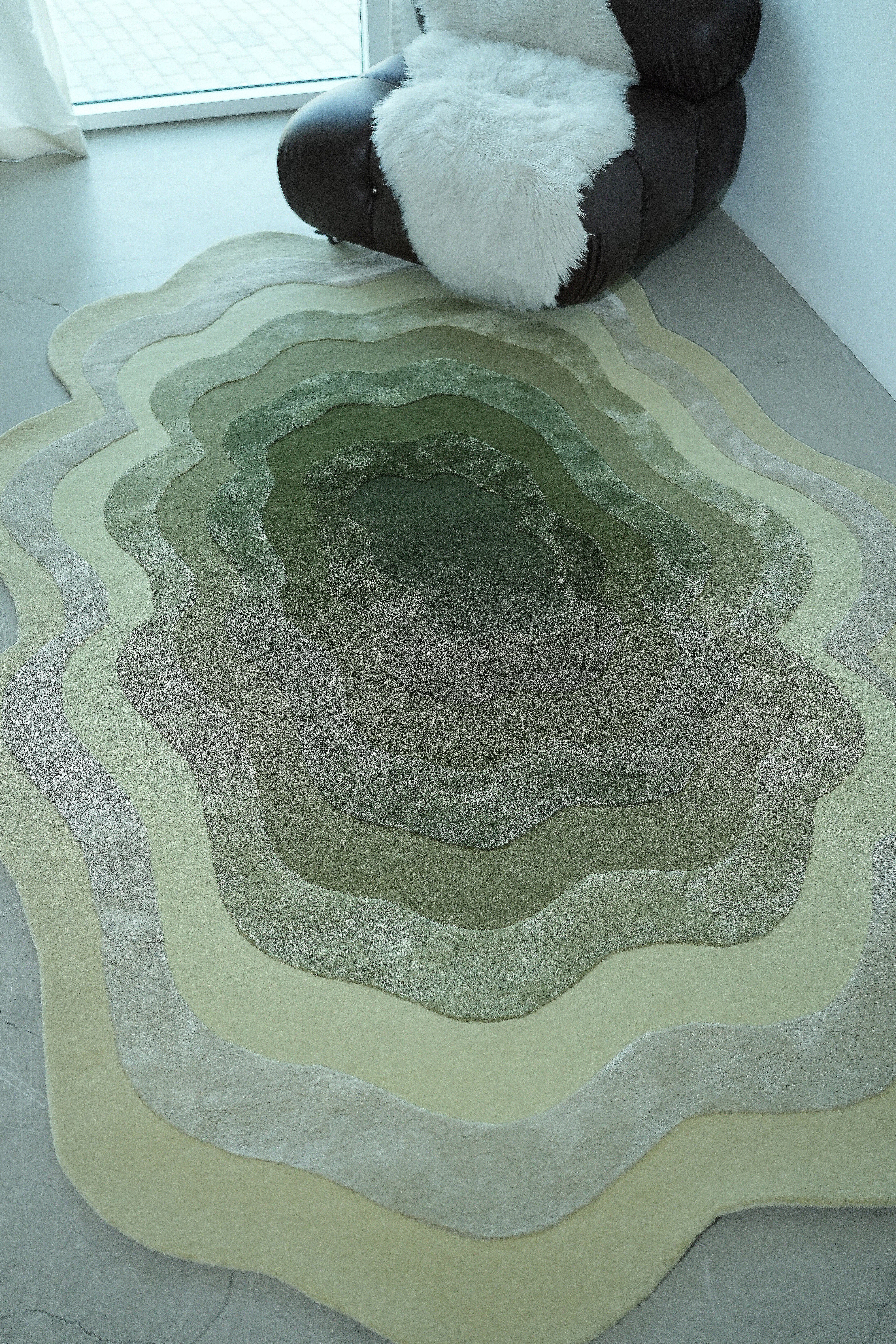The RAMSHA HOME Handmade Shape Area Rug - HTWV 164, a hand-tufted wool-viscose green abstract rug with wavy patterns, lies on a light floor beside a dark rounded chair with a white throw near a sunlit window.