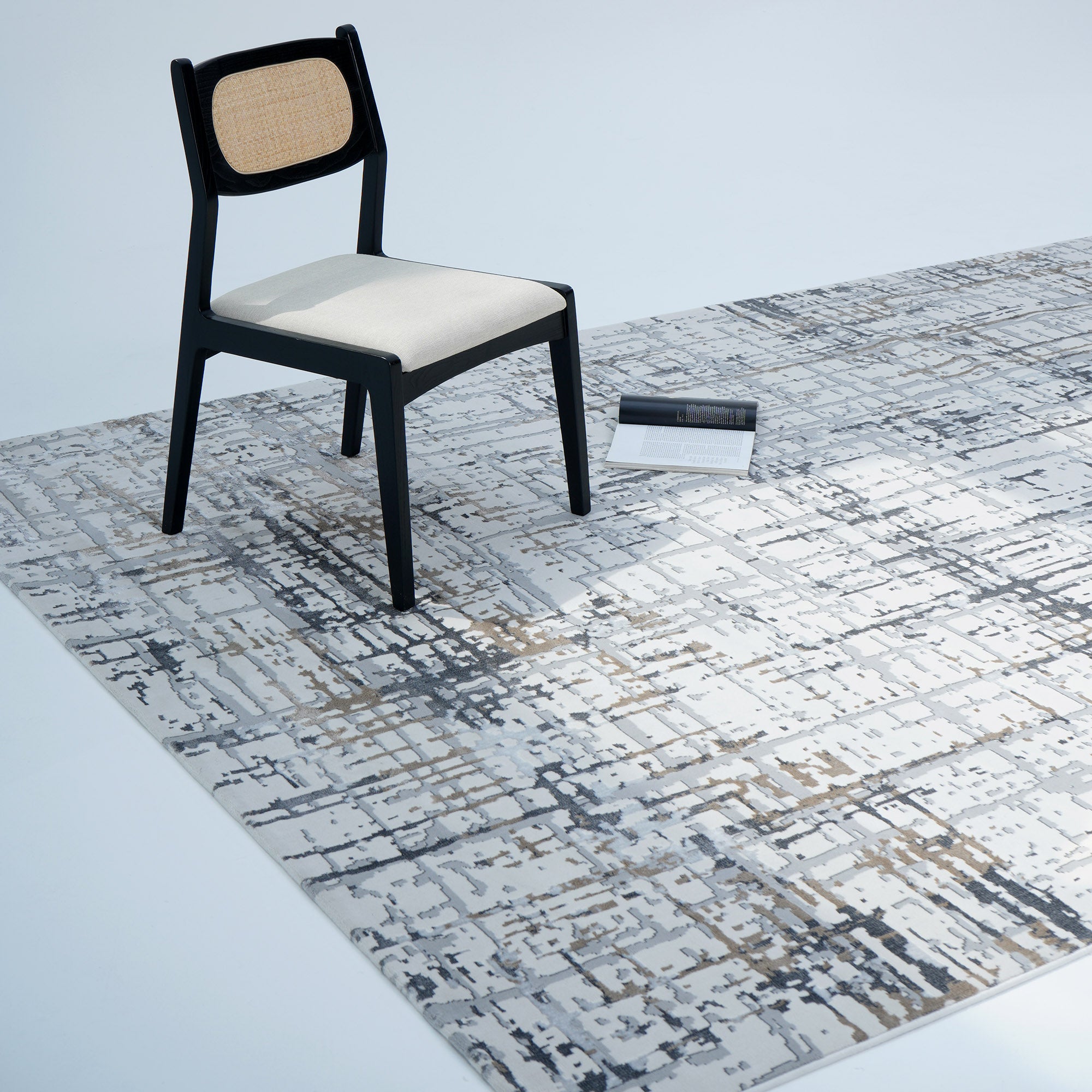 A black chair with a light seat and woven back sits on the RAMSHA HOME Cream Colored Geometry Turkish Carpet MZ24Z; a closed book is nearby, all set against a plain white background.