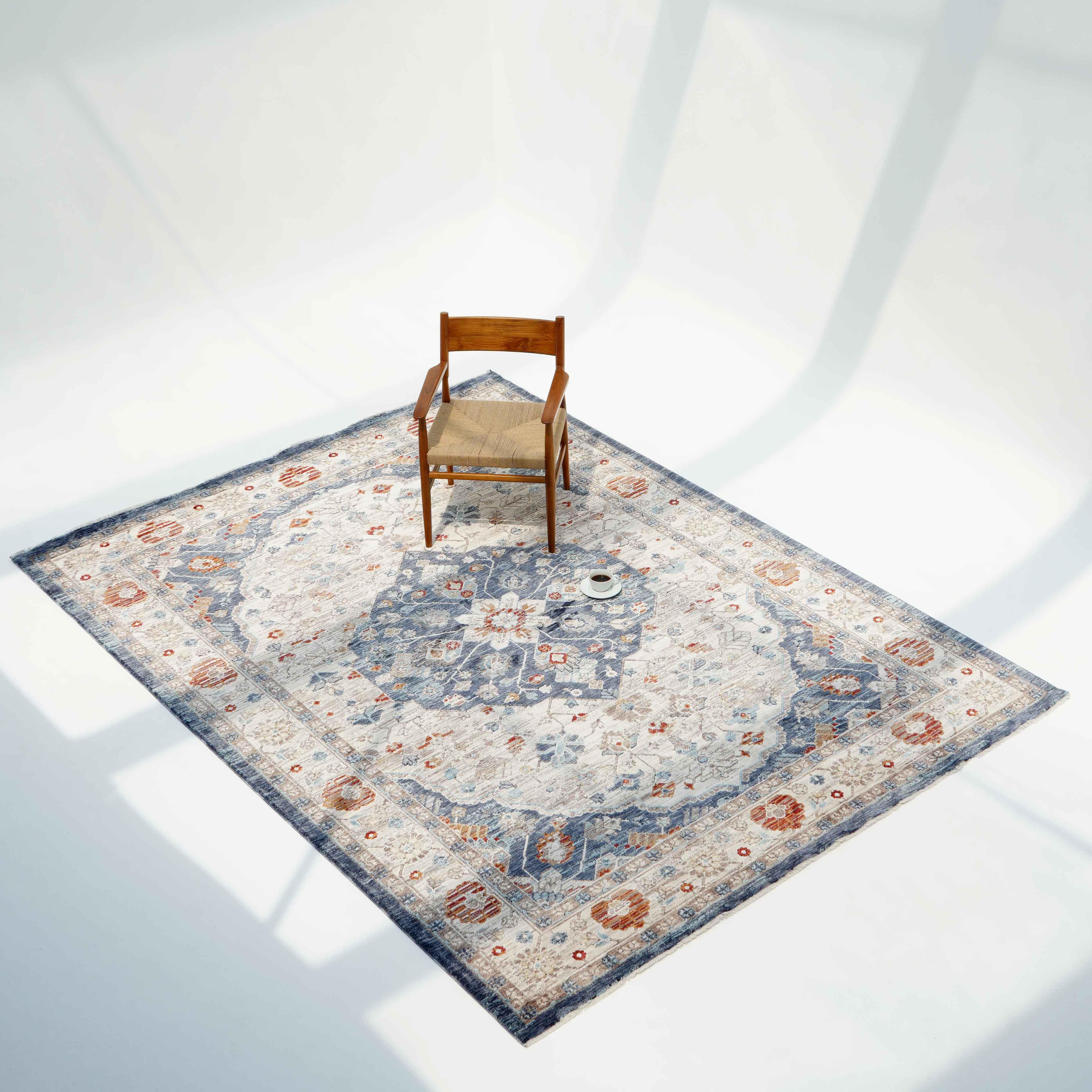 A wooden chair and a small white cup rest on the RAMSHA HOME Indoor Floor Area Rug K856B (100% Polyester Silk) featuring blue, gray, and rust medallion patterns in a softly lit room with gentle shadows on a light floor.