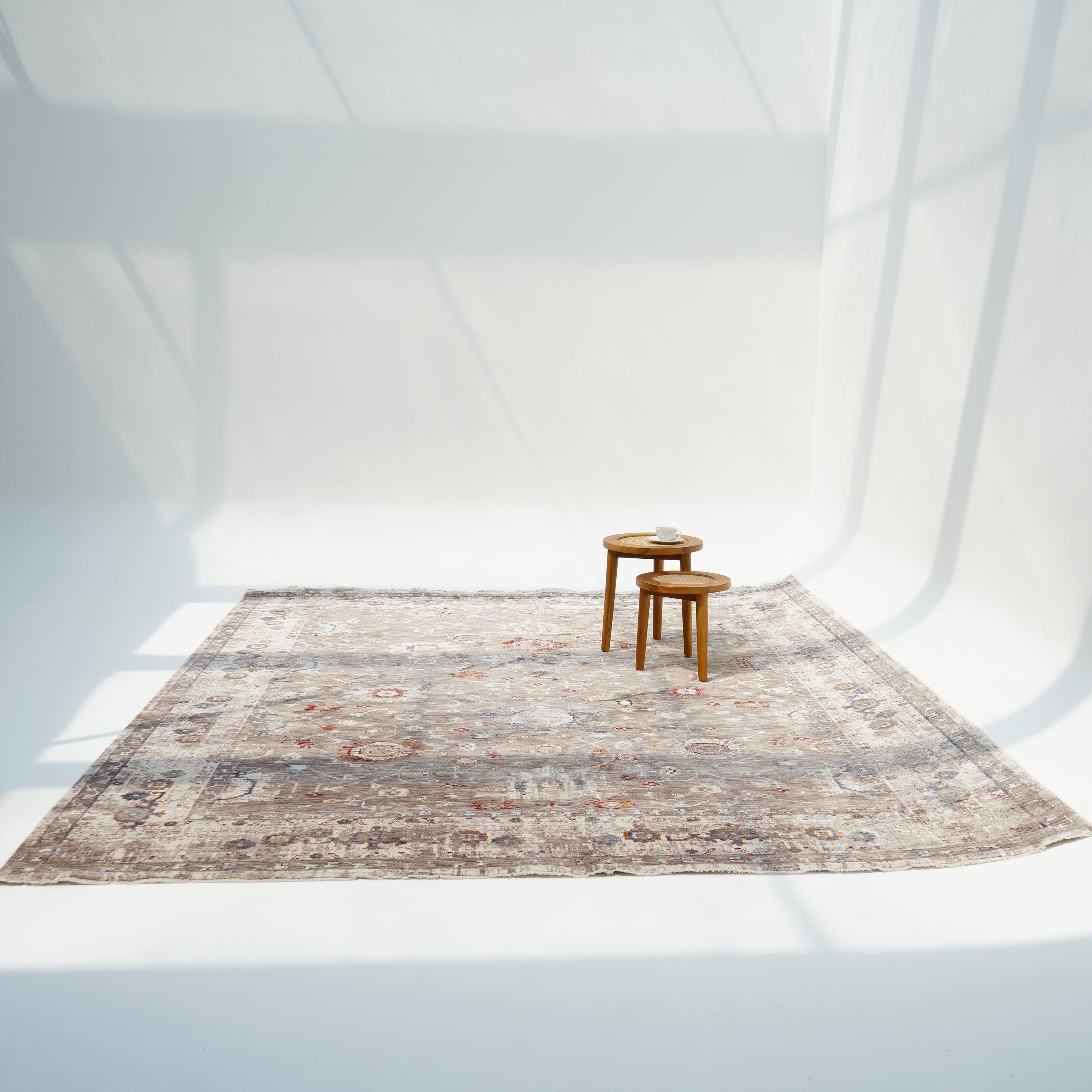 A RAMSHA HOME Beige Cream Decorative Indoor Rug K747E is placed on a white floor under soft sunlight, beside two stacked wooden stools holding a small book near the rug’s edge. Window frame shadows fall across the walls and floor.