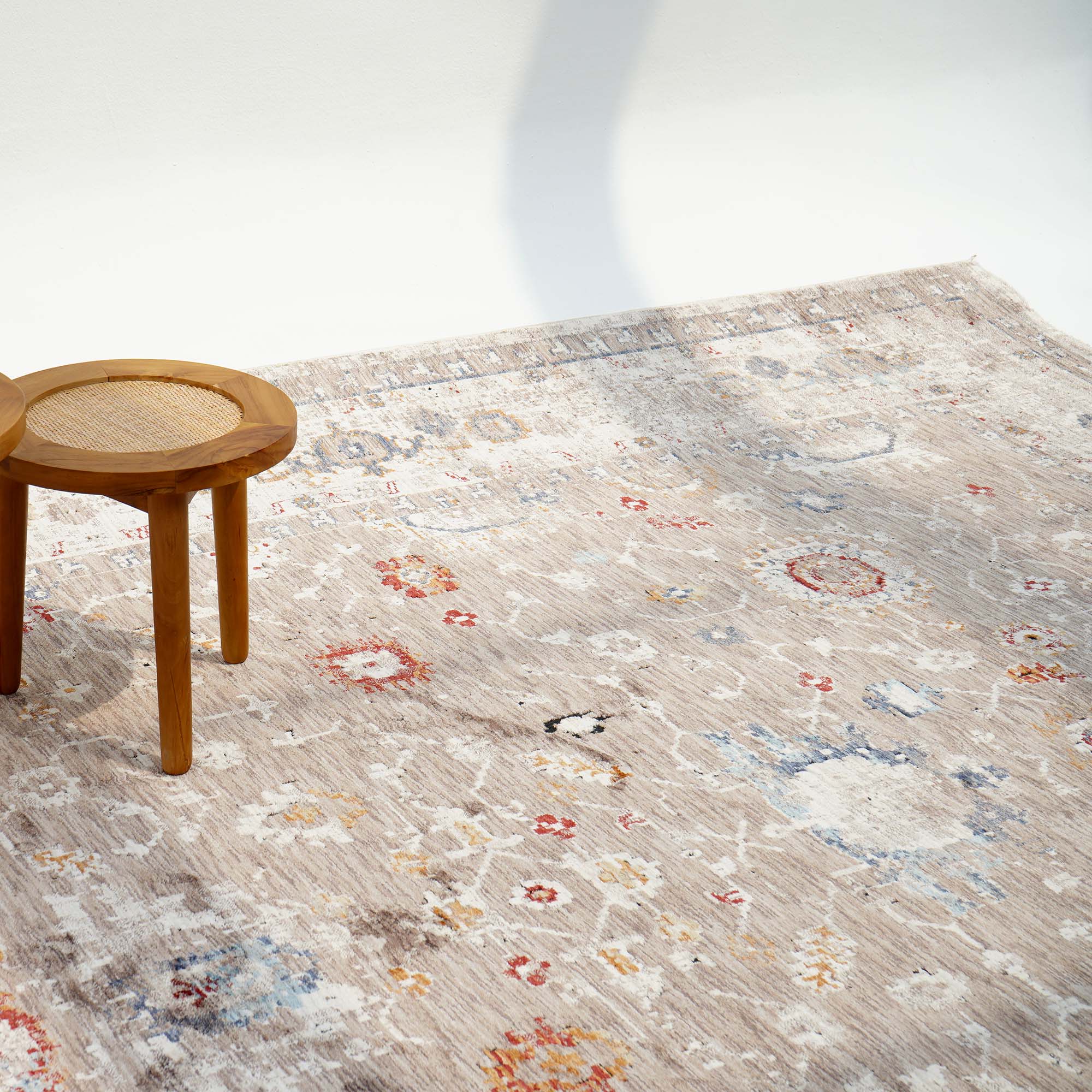 The RAMSHA HOME Beige Cream Decorative Indoor Rug K747E lies on the floor. A small round wooden stool with a woven seat sits atop the rug, while a soft shadow gently falls across the scene.