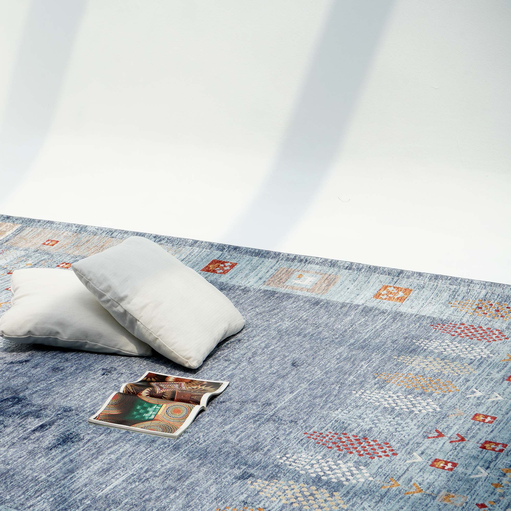 Two light gray cushions and an open magazine rest on the RAMSHA HOME Luxurious Denim Machine Made Turkey Rug - K742B, its bold geometric patterns enhancing a sunlit minimalist room where gentle shadows move across the wall.