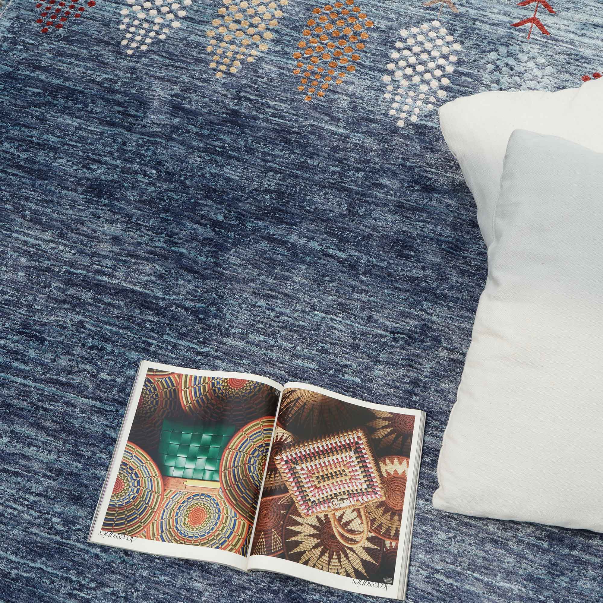 An open magazine with colorful patterns rests on the Luxurious Denim Machine Made Turkey Rug - K742B by RAMSHA HOME, a blue modern geometric rug, beside a white pillow.