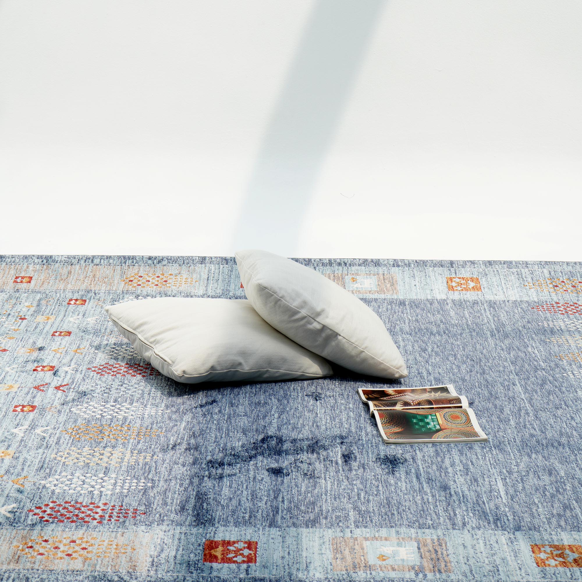Two light gray pillows sit on the RAMSHA HOME Luxurious Denim Machine Made Turkey Rug - K742B, featuring geometric blue shades. Nearby are playing cards and a magazine, all bathed in diagonal sunlight.