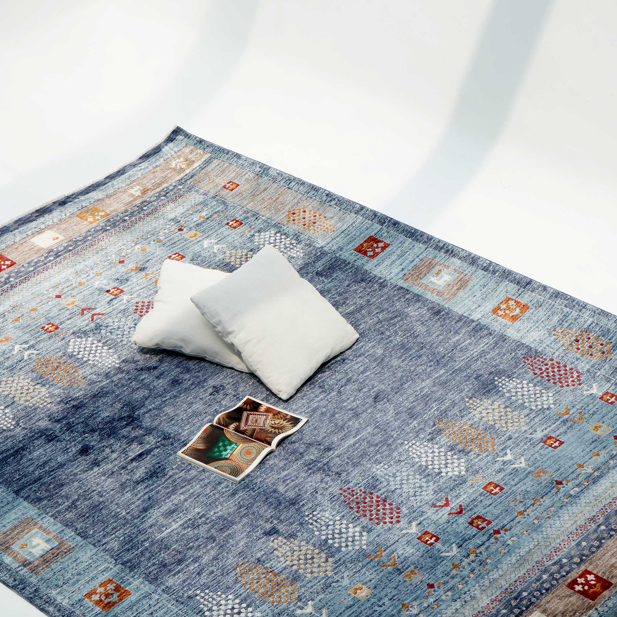 The Luxurious Denim Machine Made Turkey Rug - K742B by RAMSHA HOME features blue geometric and animal patterns, set on a light floor with two white pillows and an open magazine, enhancing the space with style and comfort.