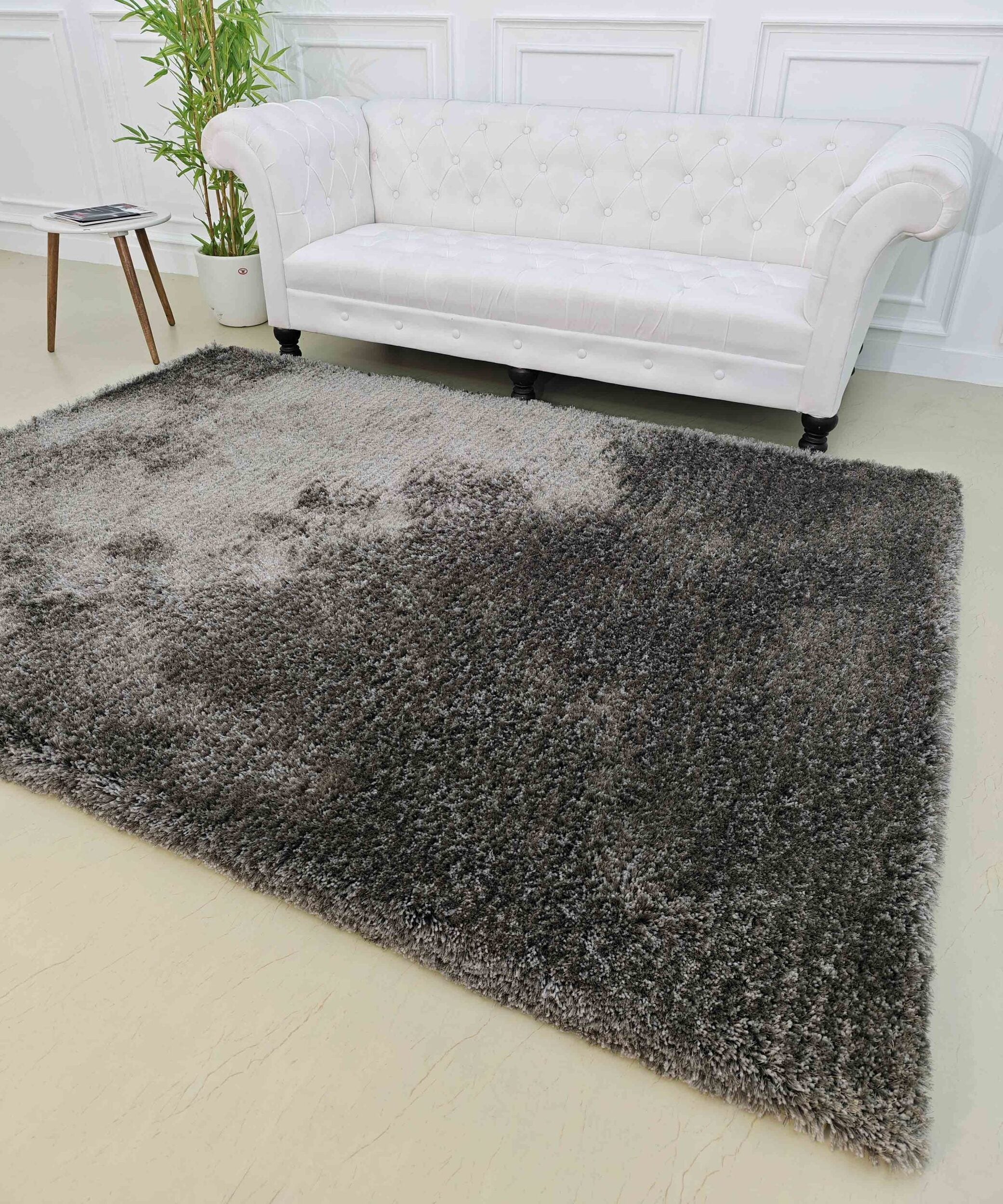 A white tufted sofa sits by a white paneled wall with a green potted plant nearby. In front is the RAMSHA HOME Grey Color Rectangle Shaped Shaggy Rug - SG 05 featuring textured brown accents on a light floor, with a small side table.