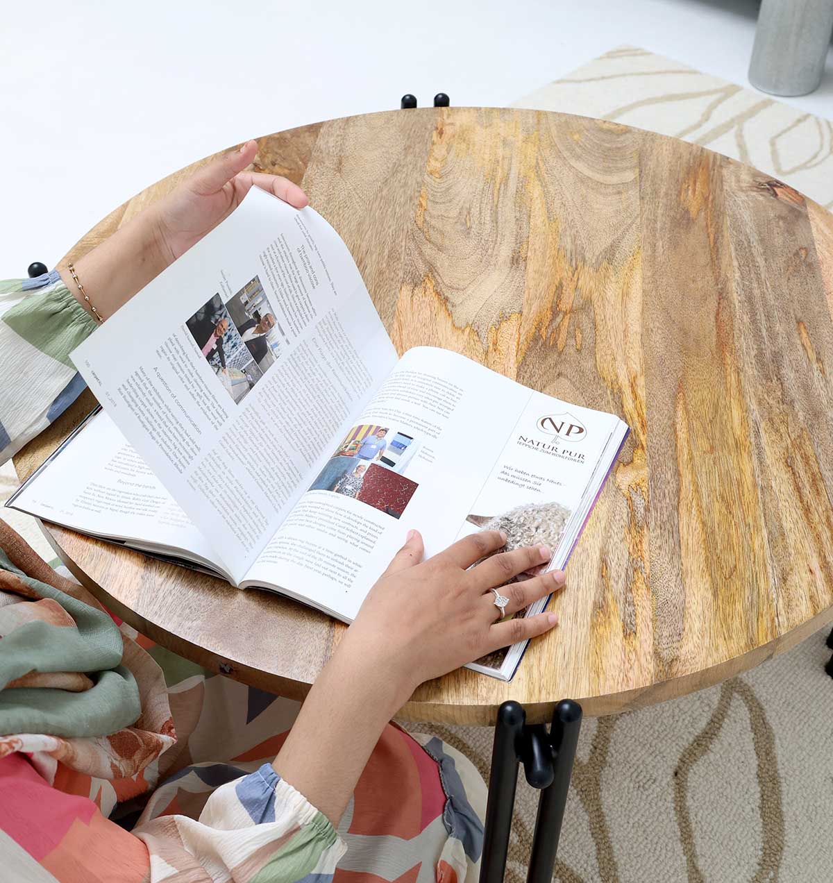 A person in colorful clothing sits at a RAMSHA HOME Modern Round Coffee Table (Solid Wood Top, Black Iron Legs GH-067), reading an open magazine. The background features part of a light, subtly patterned rug.