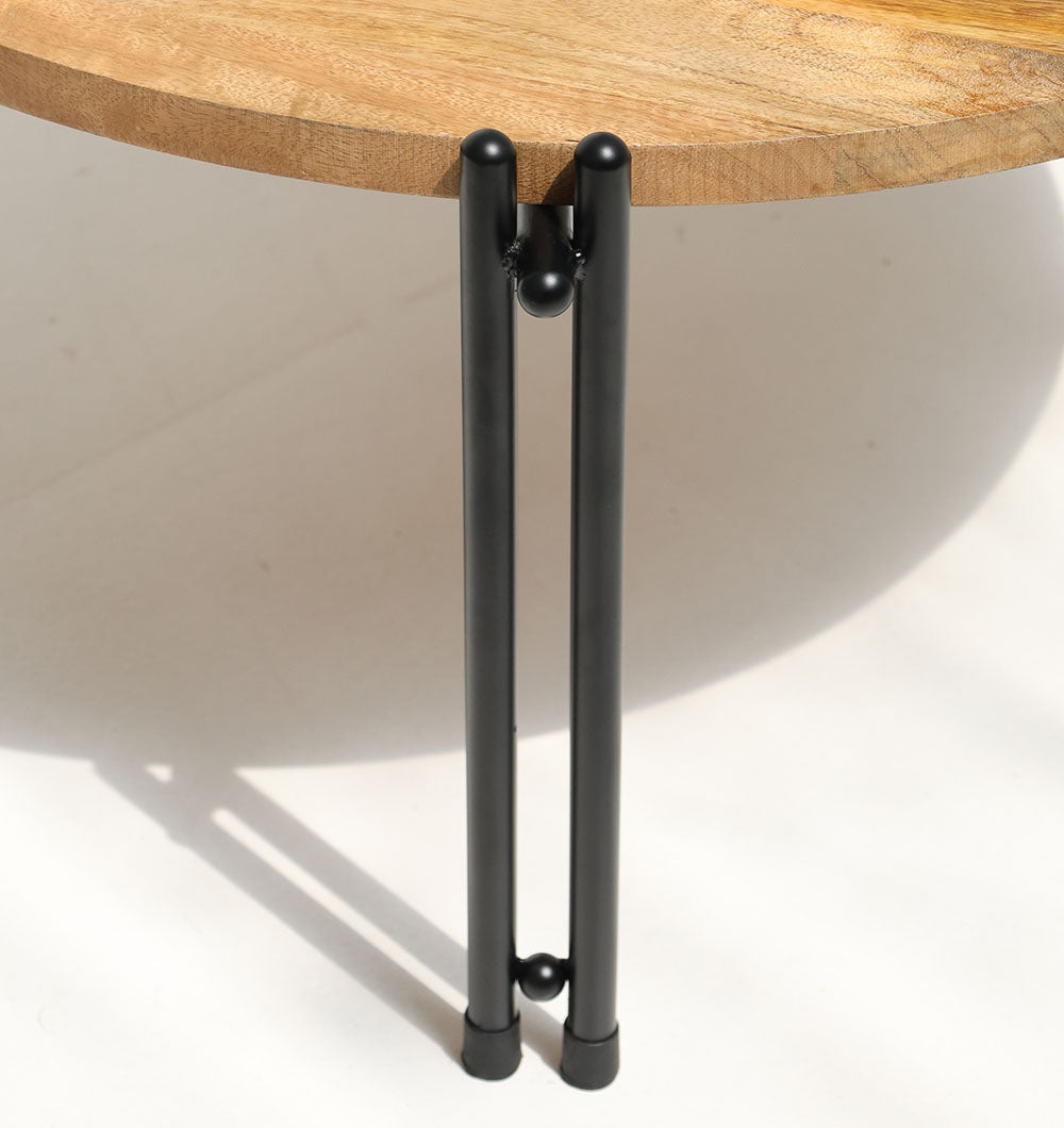 Close-up of RAMSHA HOME's Modern Round Coffee Table GH-067, featuring a solid wood top and black iron legs with parallel rods joined by spherical connectors, casting a shadow on a white surface.