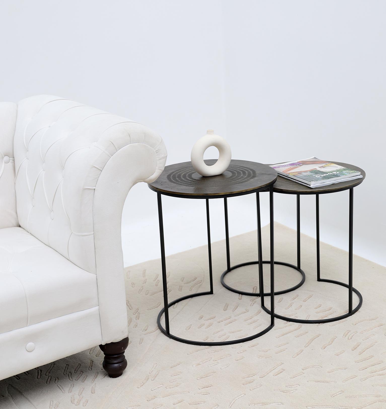 A modern living room features a white tufted sofa next to the RAMSHA HOME Set of 3 Modern Nesting Side Tables (Antique Brass Finish, Black Iron Base, GH-070); one holds a decorative white vase, another magazines, all resting on a beige patterned rug.