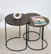 The RAMSHA HOME Set of 3 Modern Nesting Side Tables (GH-070) features antique brass tops and black iron bases. One table holds a white ceramic vase, another displays a magazine, all arranged on a light-patterned rug.