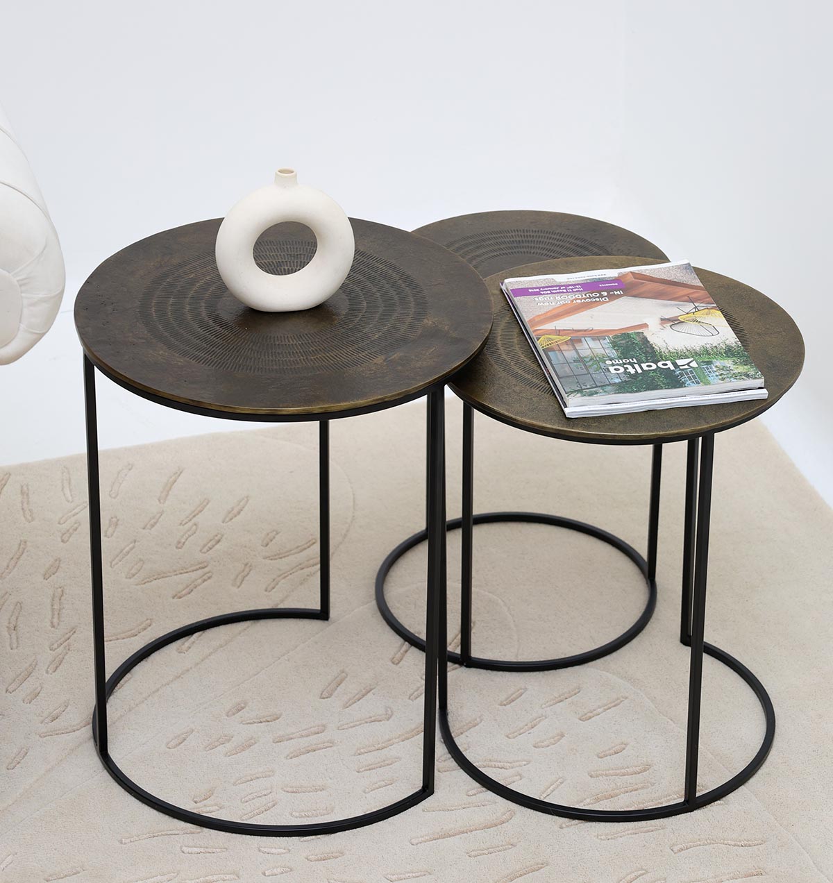 The RAMSHA HOME Set of 3 Modern Nesting Side Tables (GH-070) features antique brass tops and black iron bases. One table holds a white ceramic vase, another displays a magazine, all arranged on a light-patterned rug.