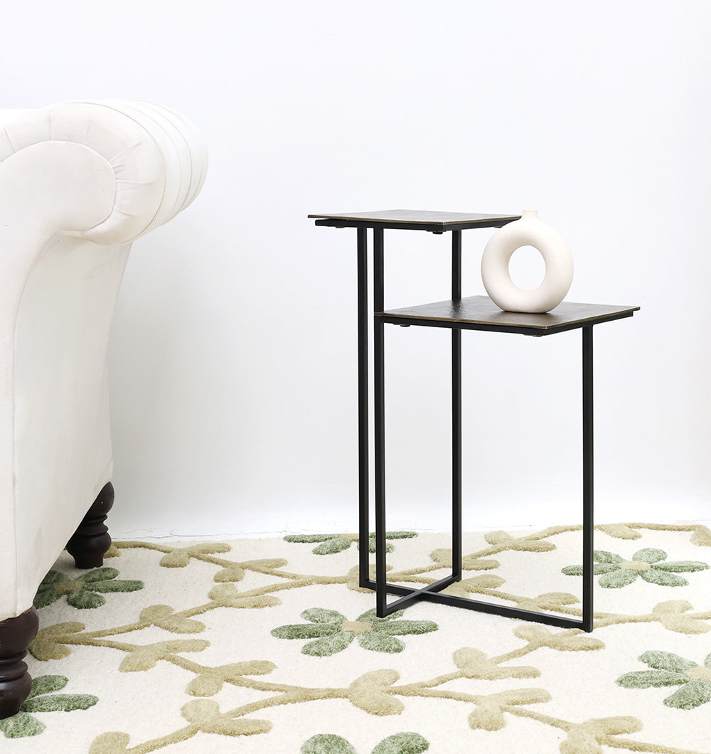 A RAMSHA HOME Modern Dual-Tier Side Table – Antique Brass Finish with Black Iron Base (GH-075-A) stands by a white sofa, holding a round vase on its two tiers atop a cream rug with a green floral pattern.