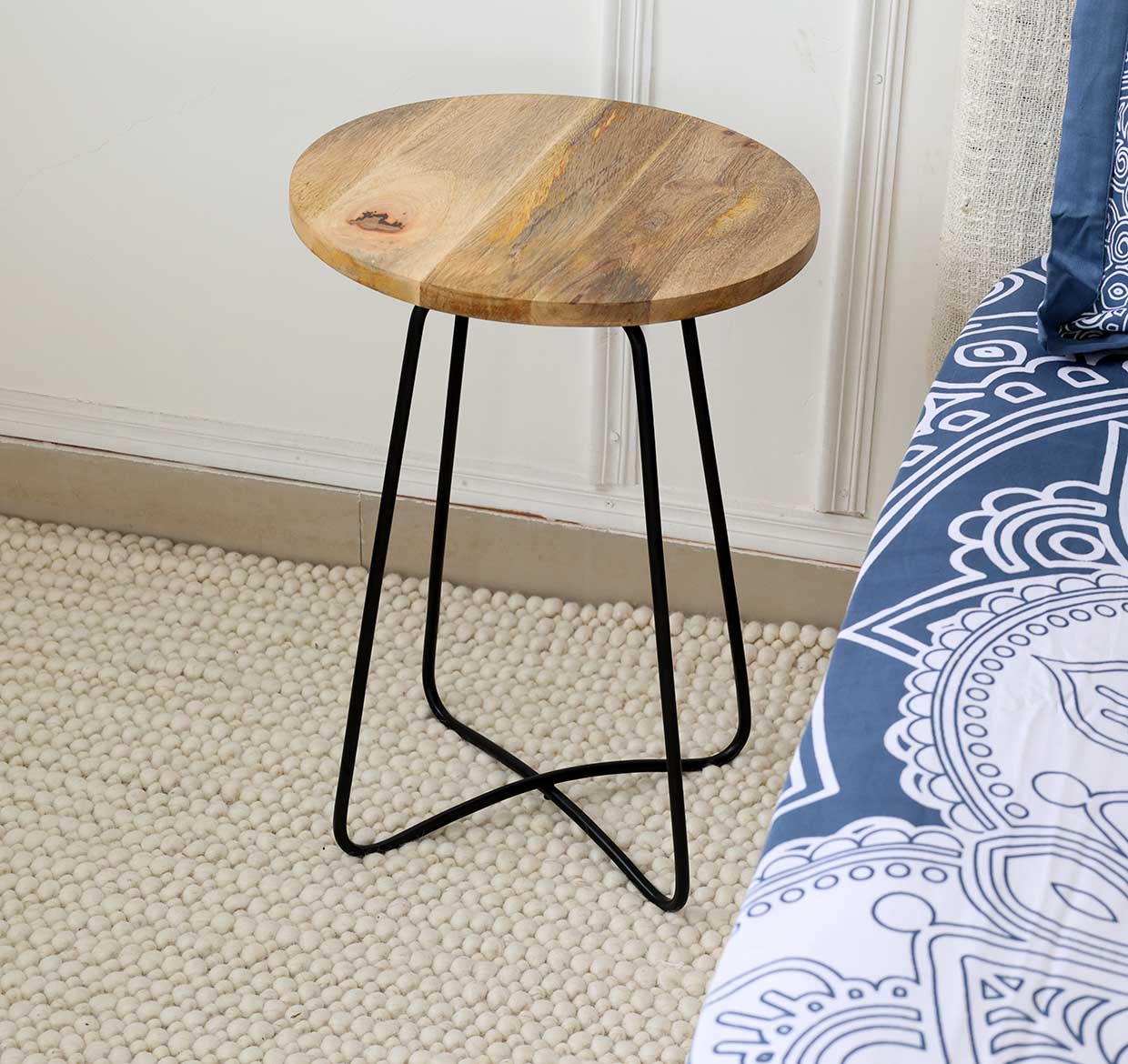 A RAMSHA HOME Modern Round Side Table – Natural & Black Powder Finish with Black Iron Base (GH-075-C) sits beside a bed with a blue and white patterned comforter on a textured beige carpet.
