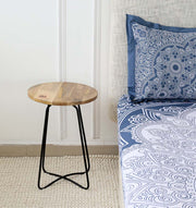 The RAMSHA HOME Modern Round Side Table GH-075-C, featuring a natural top and black iron base, stands by the bed with blue and white mandala bedding. A white textured wall and light patterned rug enhance the cozy, stylish room.
