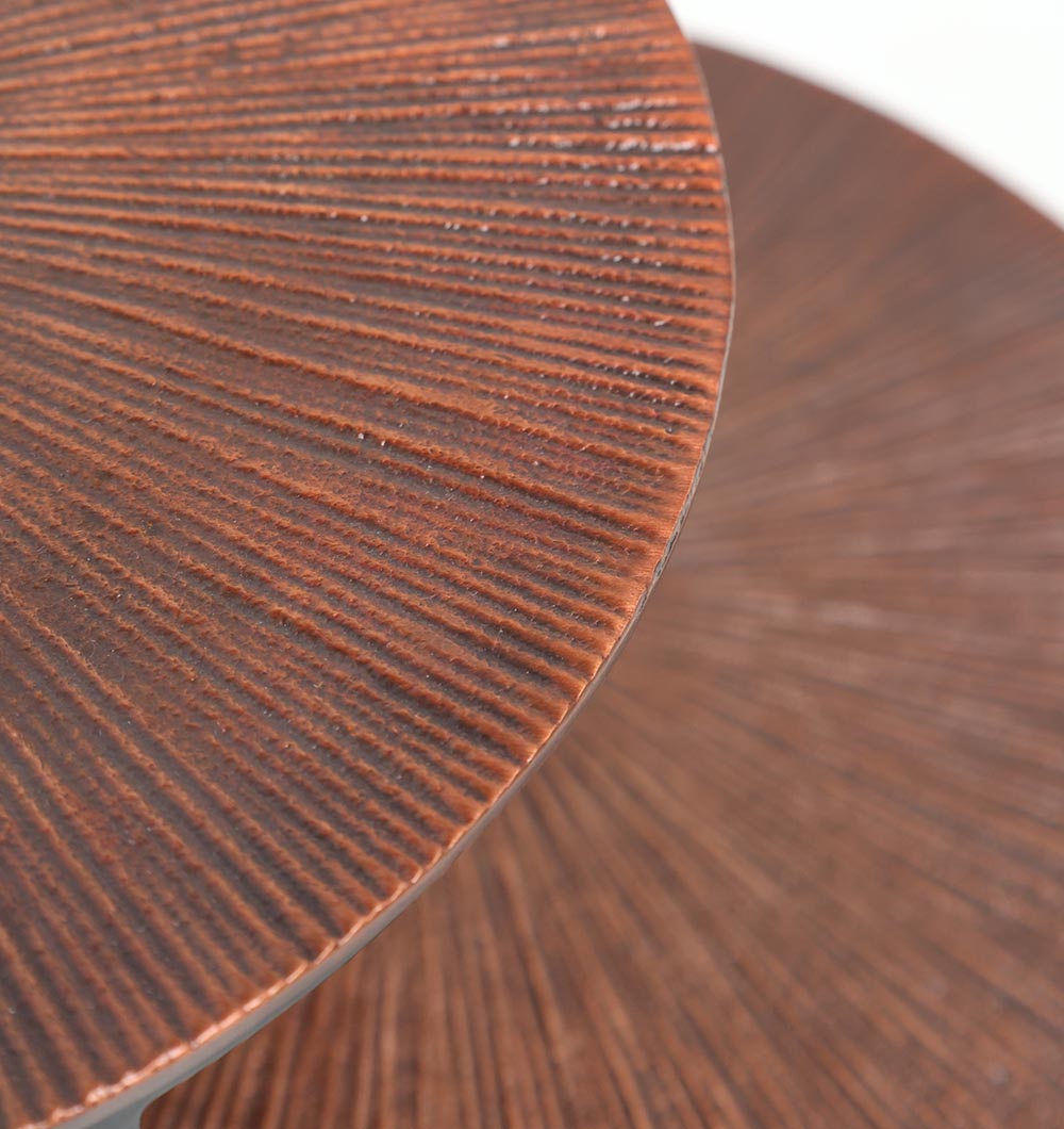 Close-up of the RAMSHA HOME Modern Dual-Tier Side Round Table (GH-075-C) shows two copper antique finish surfaces with fine radiating lines resembling sunburst wood grain, slightly overlapping on a black iron base; background blurred.