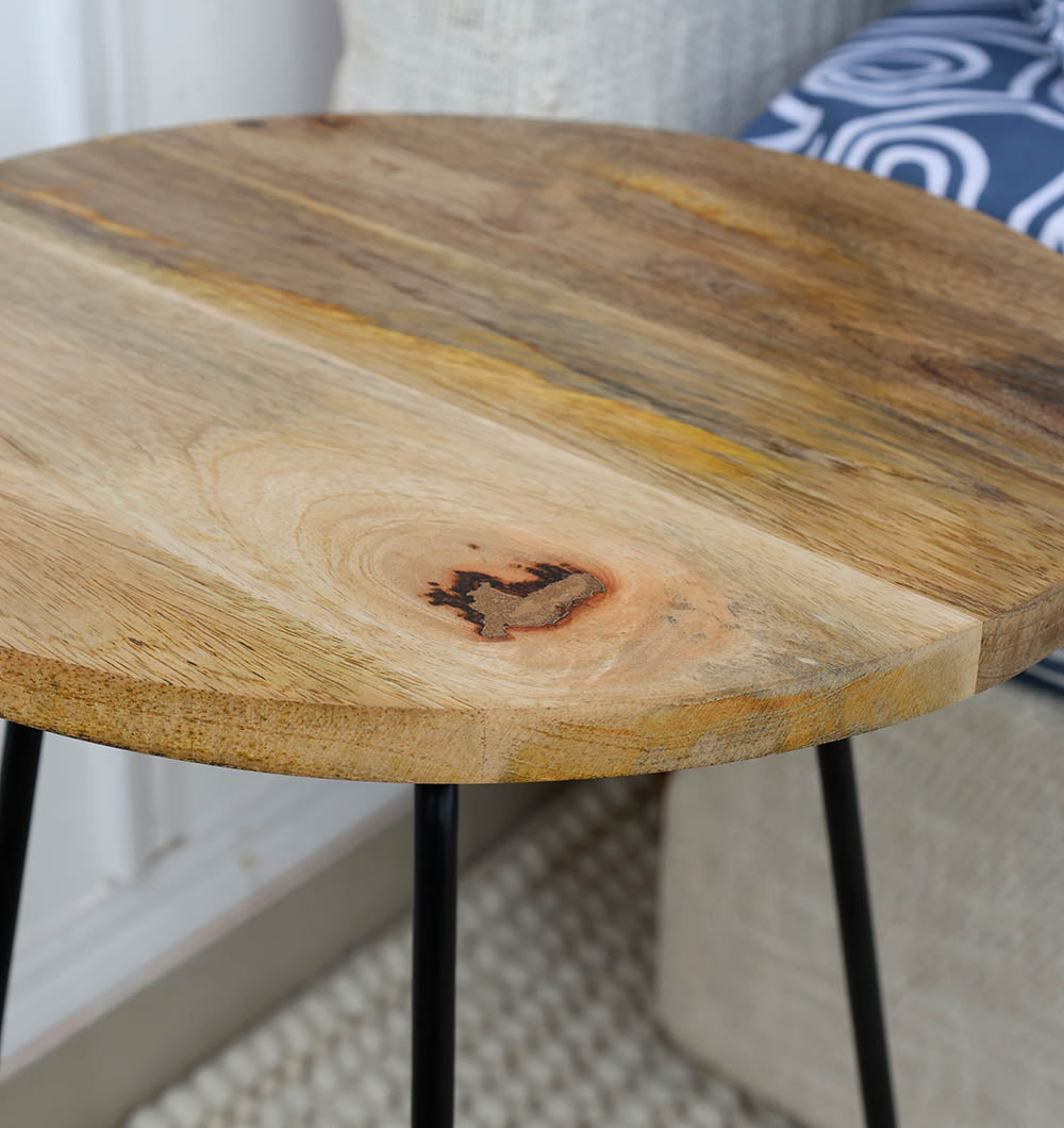 The RAMSHA HOME Modern Round Side Table (GH-075-C) features a natural wood top with visible grain and black iron base, finished in black powder. It's shown next to a patterned cushion and a textured rug.