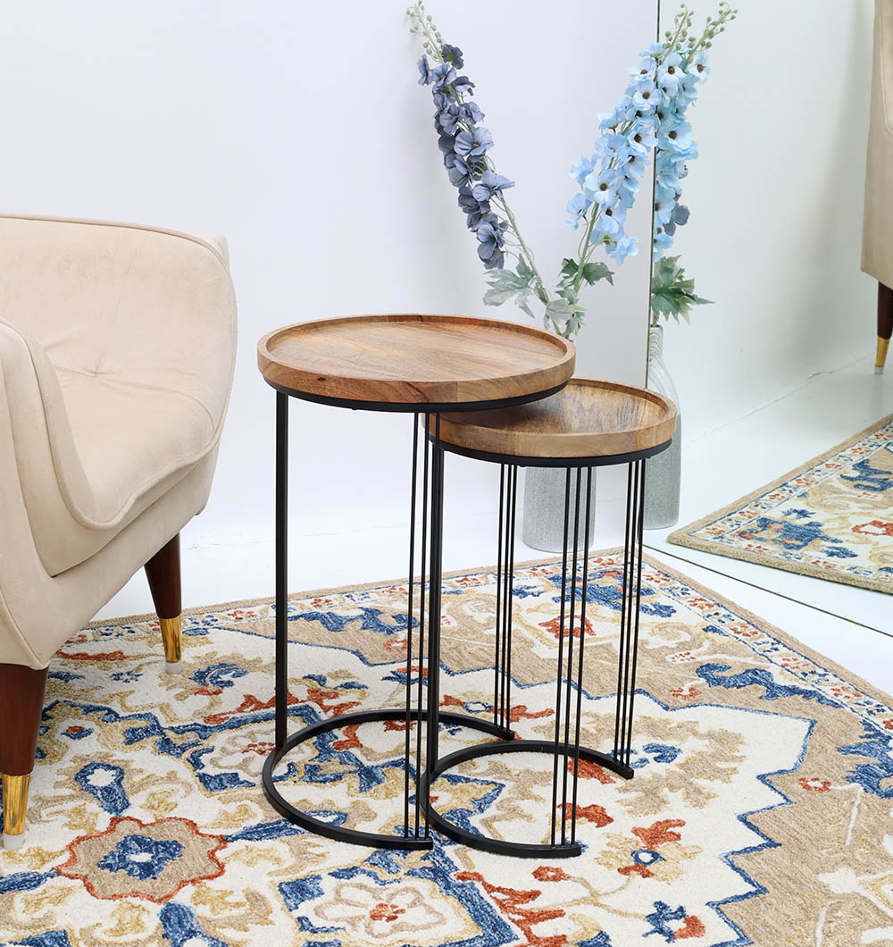 A RAMSHA HOME Set of 2 Wooden Nesting Side Tables with solid wood tops and black iron bases sits on a patterned rug beside a beige armchair, with a mirror and blue flowers in the background.